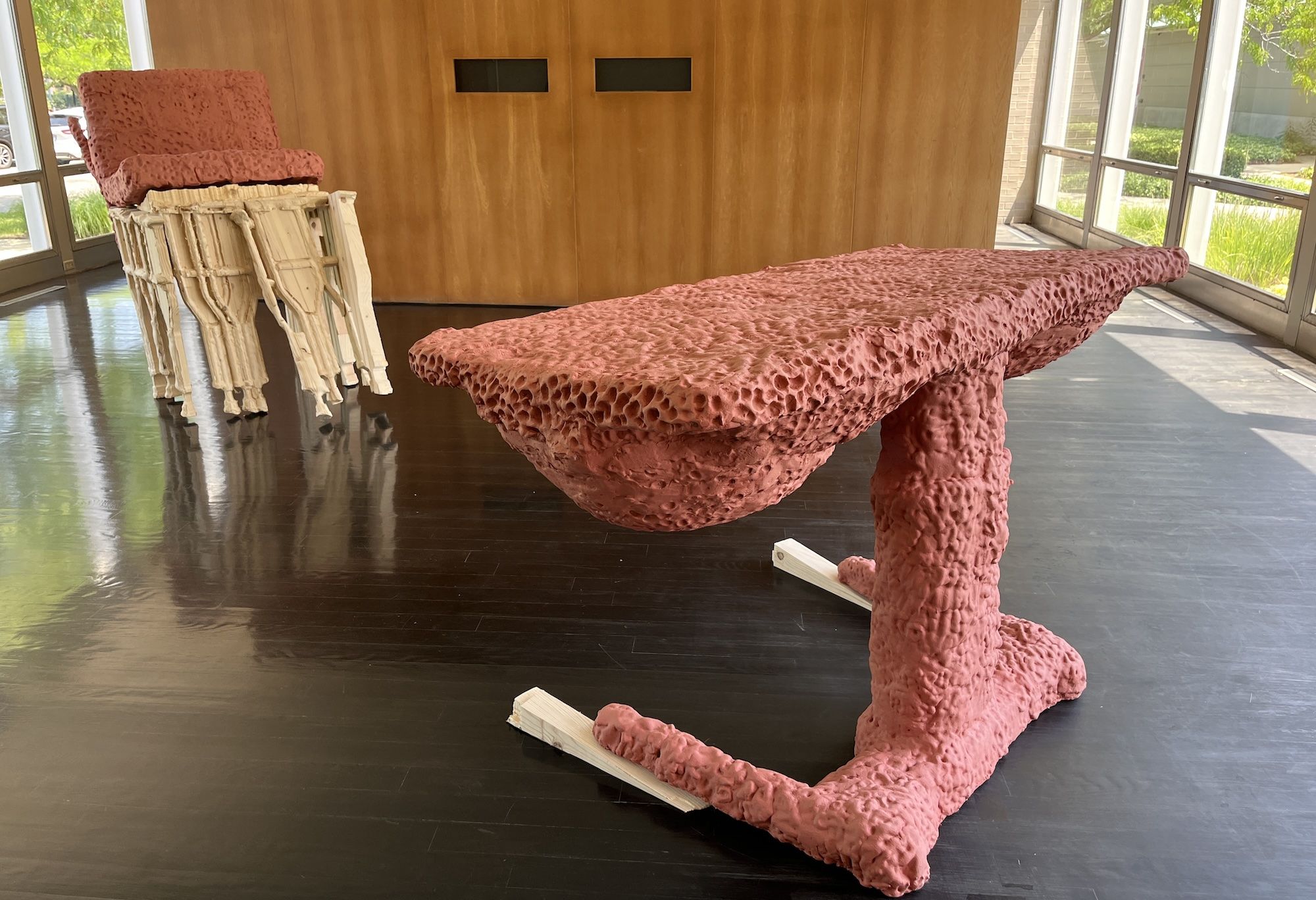 Interior room with a long pink bubbly-textured sofa and a matching pink chair on a stack of white chairs. A wood-paneled wall with two black openings and glass walls looking out on greenery.