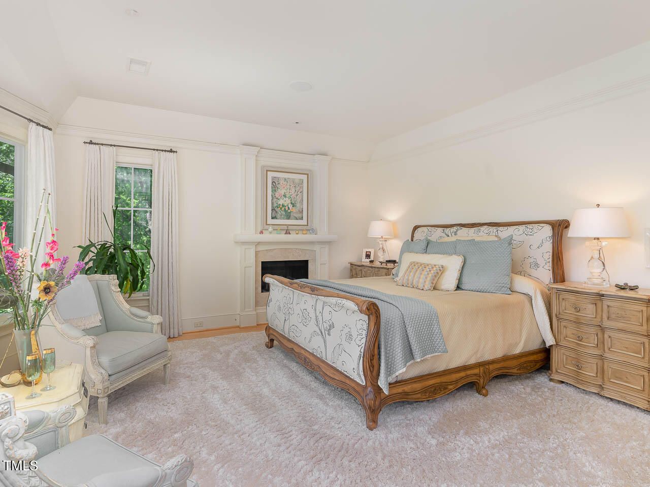 Bright bedroom with white walls, large bed with wood and floral upholstery, beige and blue bedding, two wooden nightstands with lamps, a white fireplace, and armchairs with floral decorations.