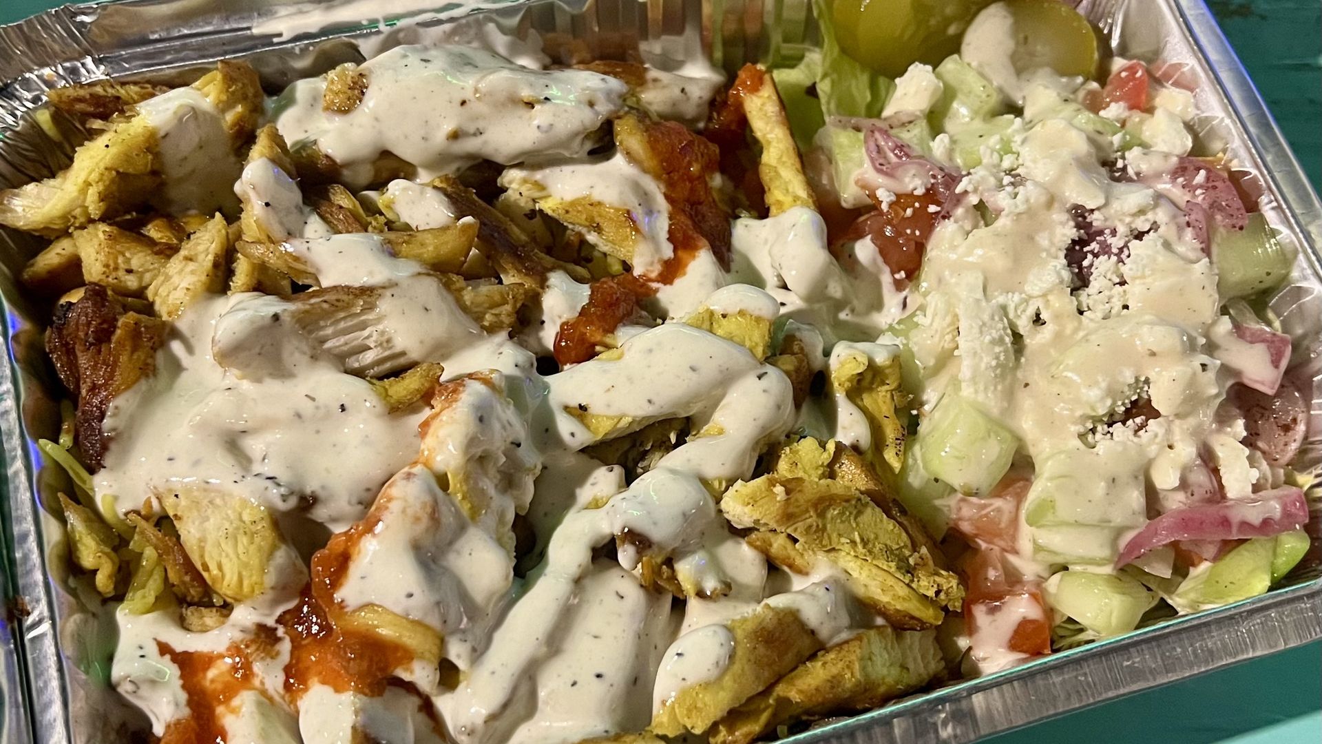 Foil tray with grilled chicken strips covered in white sauce, some red sauce, and a side salad of cucumbers, tomatoes, onions, lettuce, olives, and white crumbled cheese, all lightly dressed.