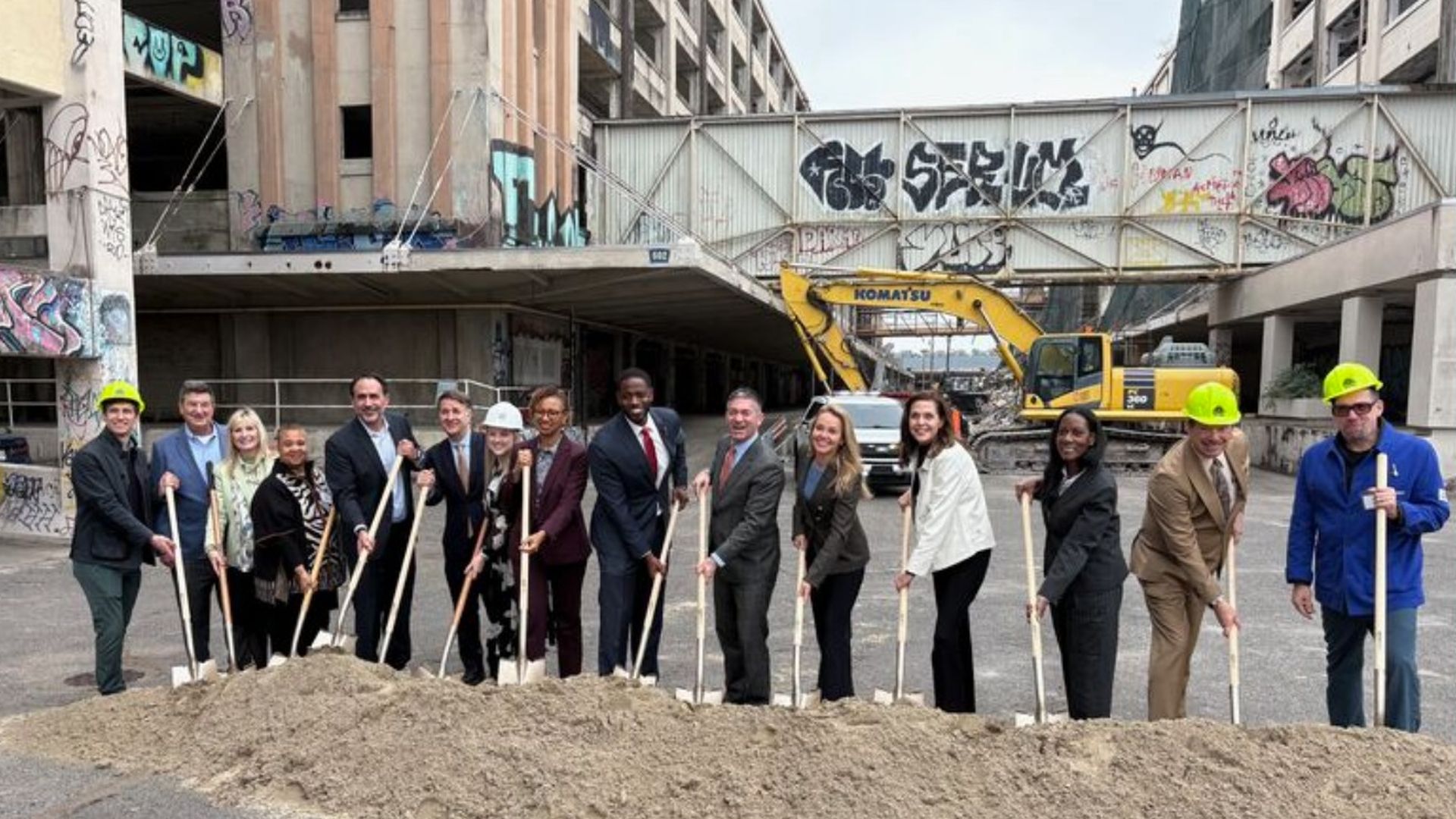Group of 15 people in business and construction attire holding shovels in front of a dirt pile at a graffiti-covered urban construction site with a yellow excavator in the background.