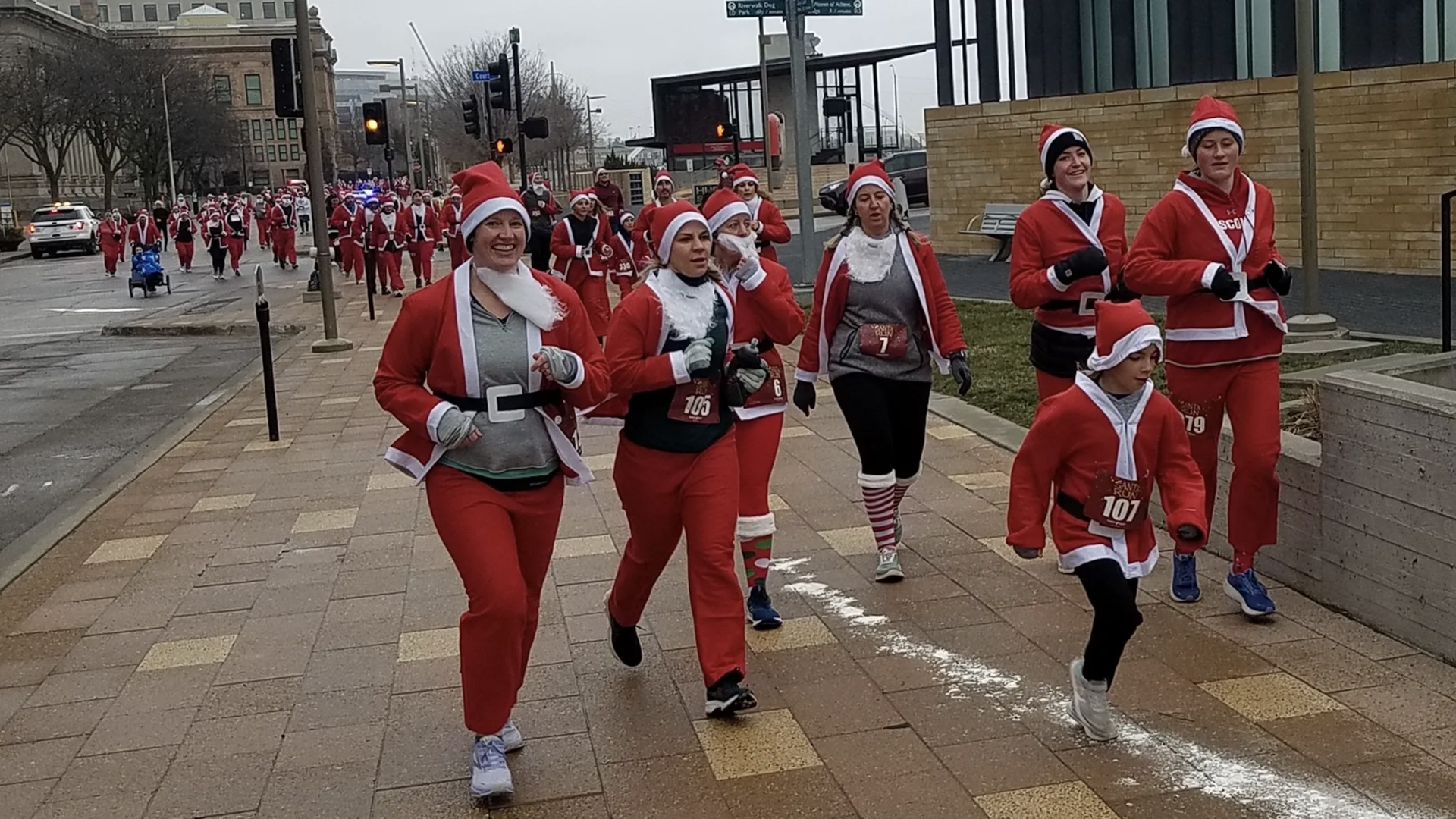 A group of people running or walking on a city sidewalk dressed in Santa Claus costumes with red suits and hats, participating in a festive outdoor race during daytime.