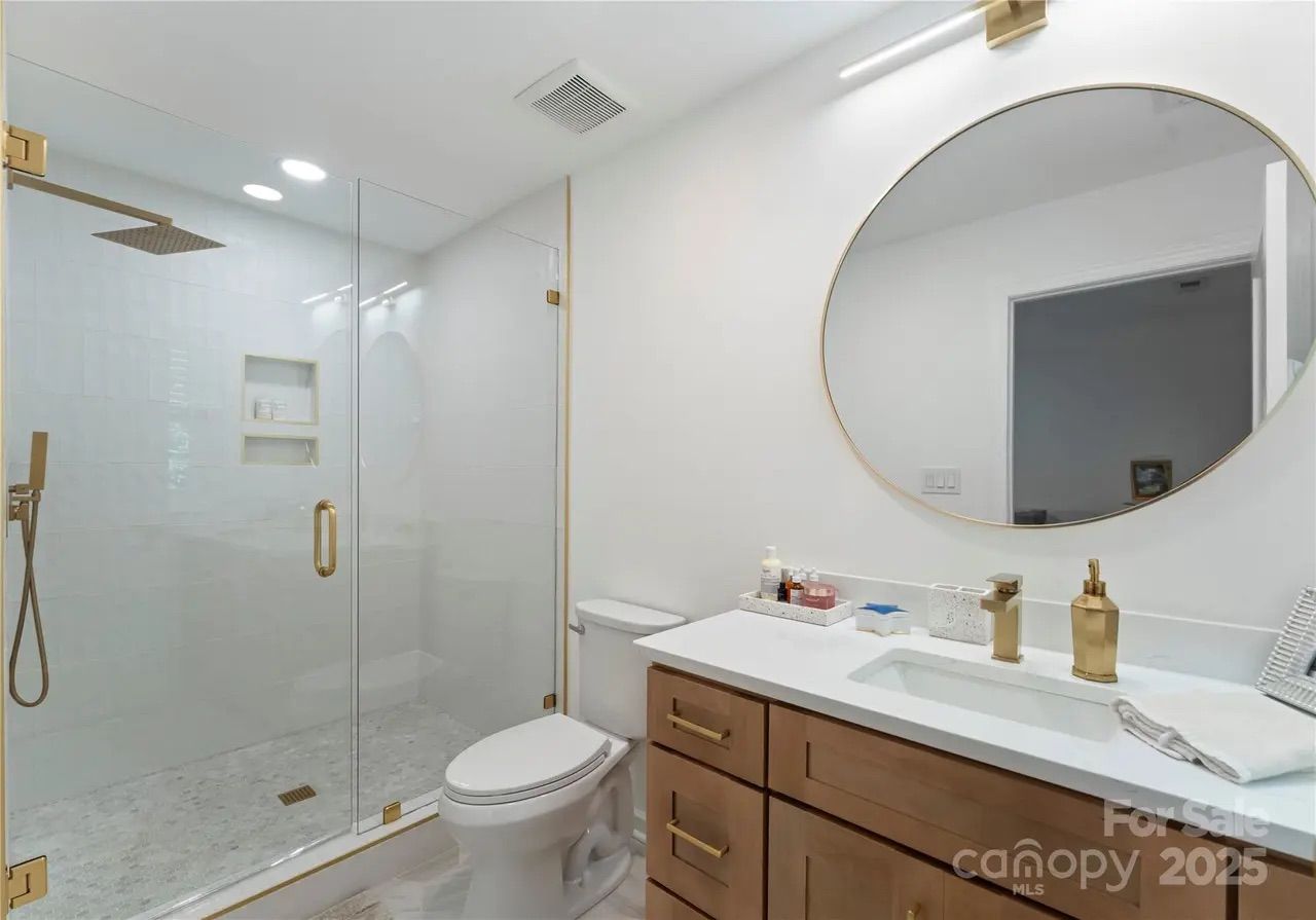 Modern bathroom with glass shower, gold fixtures, white countertop, wooden cabinets, large round mirror, and a toilet, featuring white walls and marble-patterned floor.