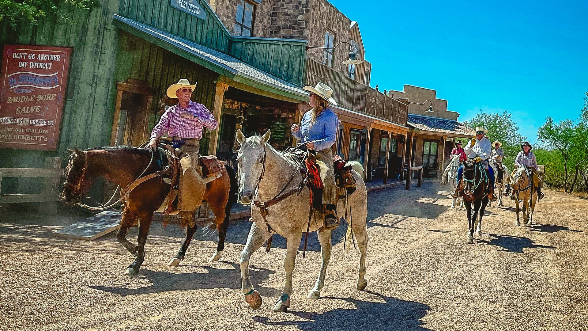 Blue sky over a western main street with green storefronts and a stone building. Cowboys ride horses along the dusty road past signs for the Post Office and Wells Fargo.