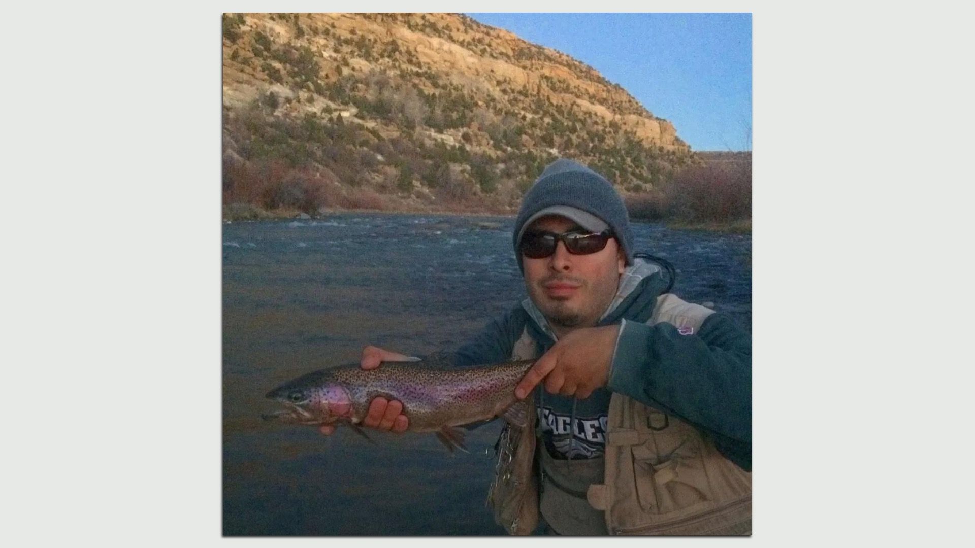 Steven Fernandez in a gray beanie and sunglasses holds a large rainbow trout over a river.