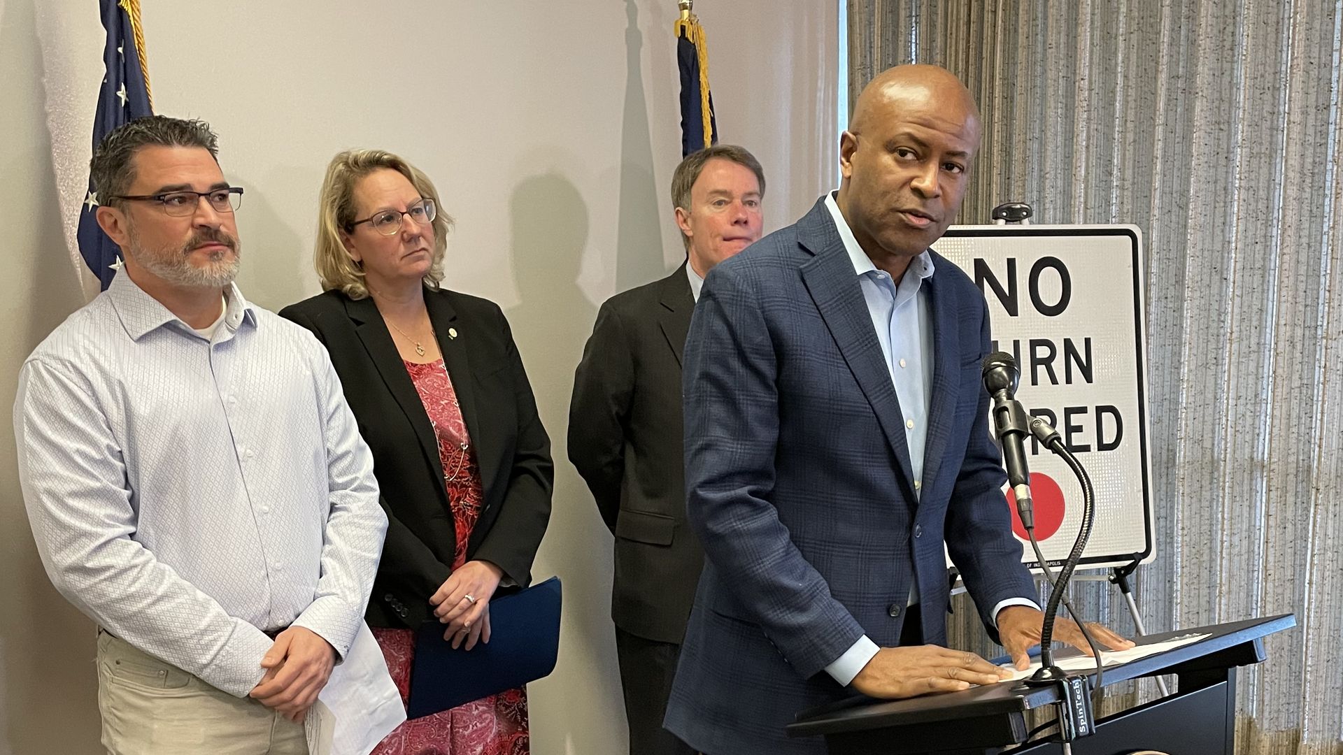City-County Council President Vop Osili speaks into a microphone while councilors Zach Adamson and Kristin Jones and Mayor Joe Hogsett look on from behind.