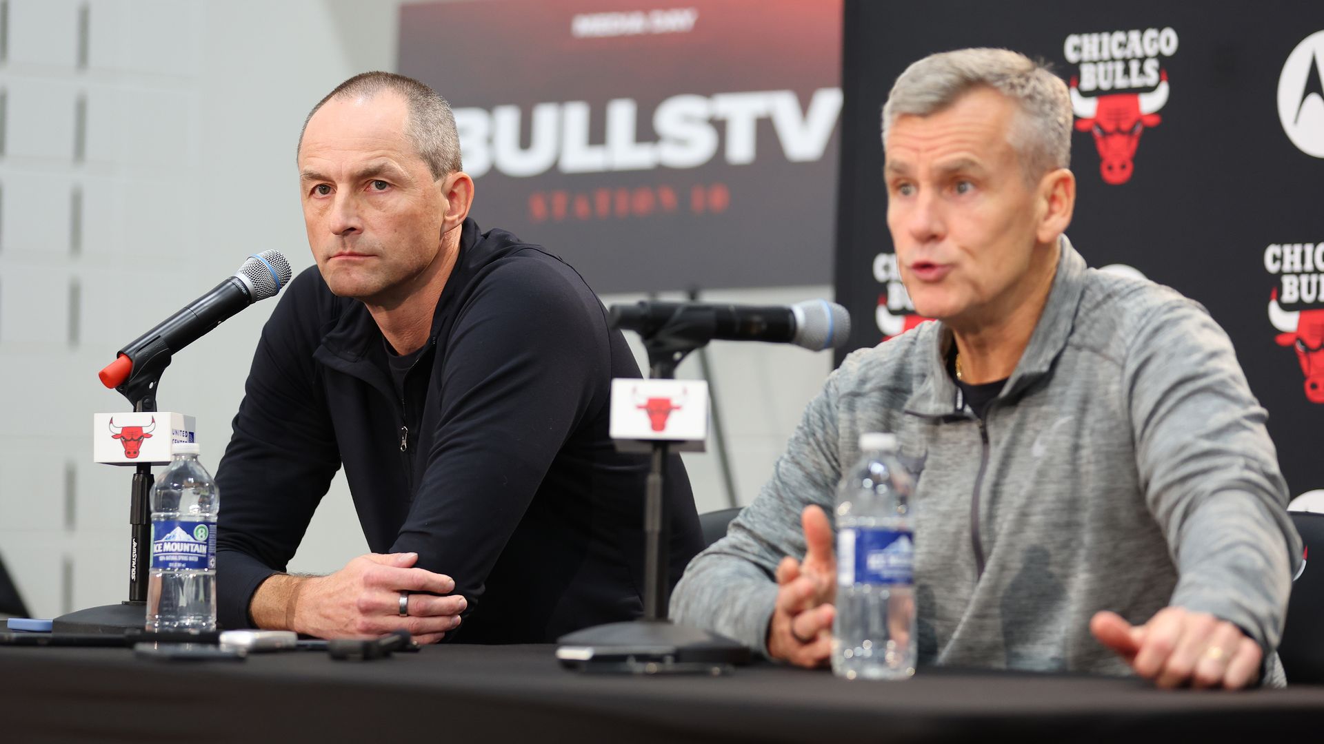 Photo of two men at a podium in front of microphones talking to media 