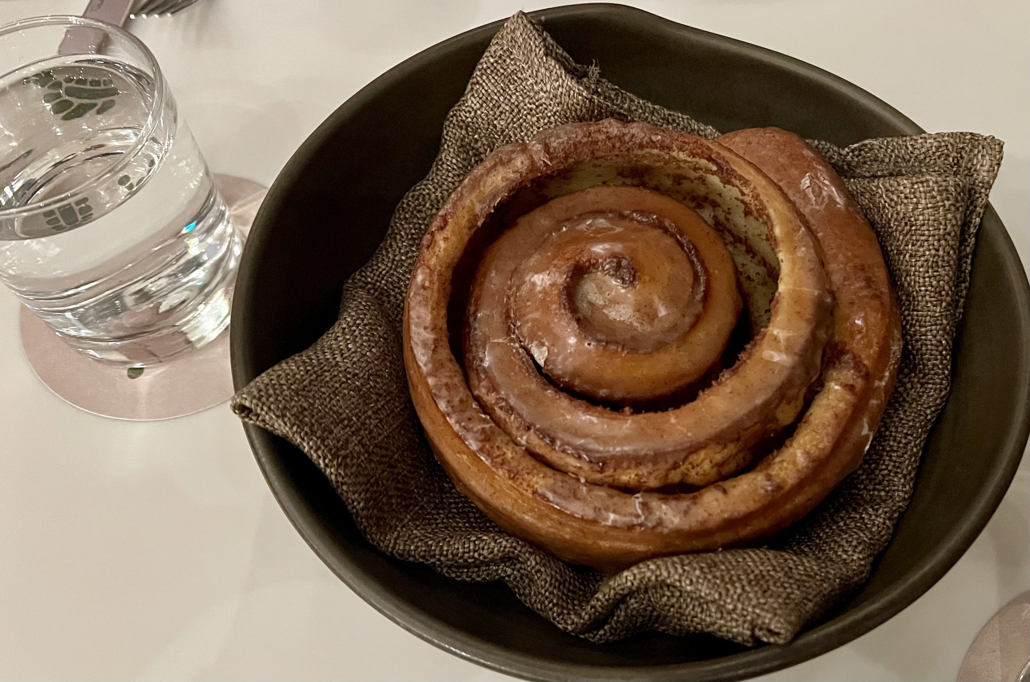 Glazed cinnamon roll on a brown cloth napkin inside a dark bowl, with a clear glass of water on a coaster beside it on a white surface.