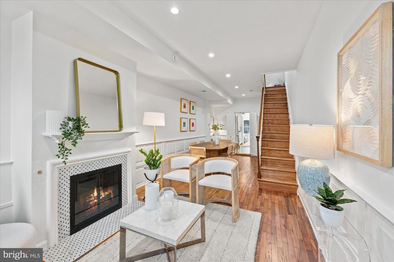 Bright living room with white walls and wood floors. A fireplace with a gold-framed mirror above, white tile surround and greenery. Two light wood chairs, marble coffee table, rug, stairs to the right, dining area in back.