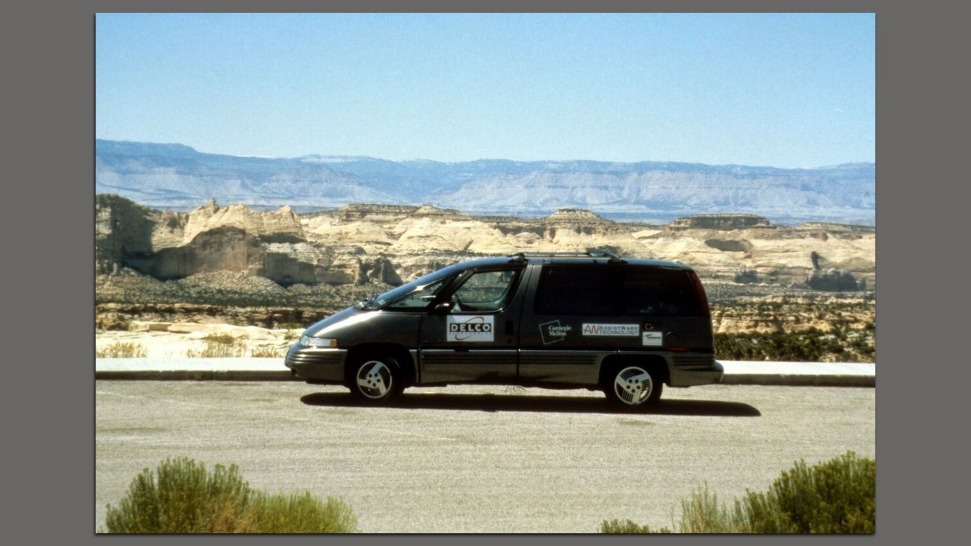 A dark minivan with sponsor decals, including DELCO, sits on a desert roadside. Pale rock canyons and distant blue mountains rise under a clear blue sky.