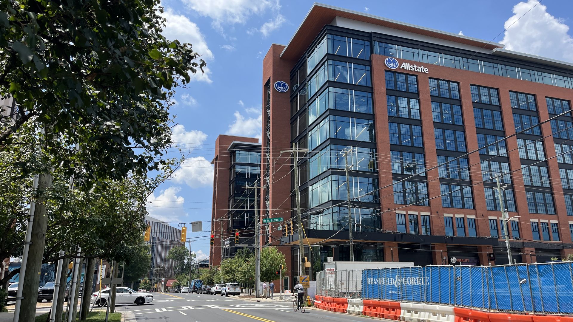Modern brick-and-glass office building with the Allstate logo on a city street; construction barriers along the curb, trees on the left, blue sky with clouds, and cars and pedestrians at the crosswalk.