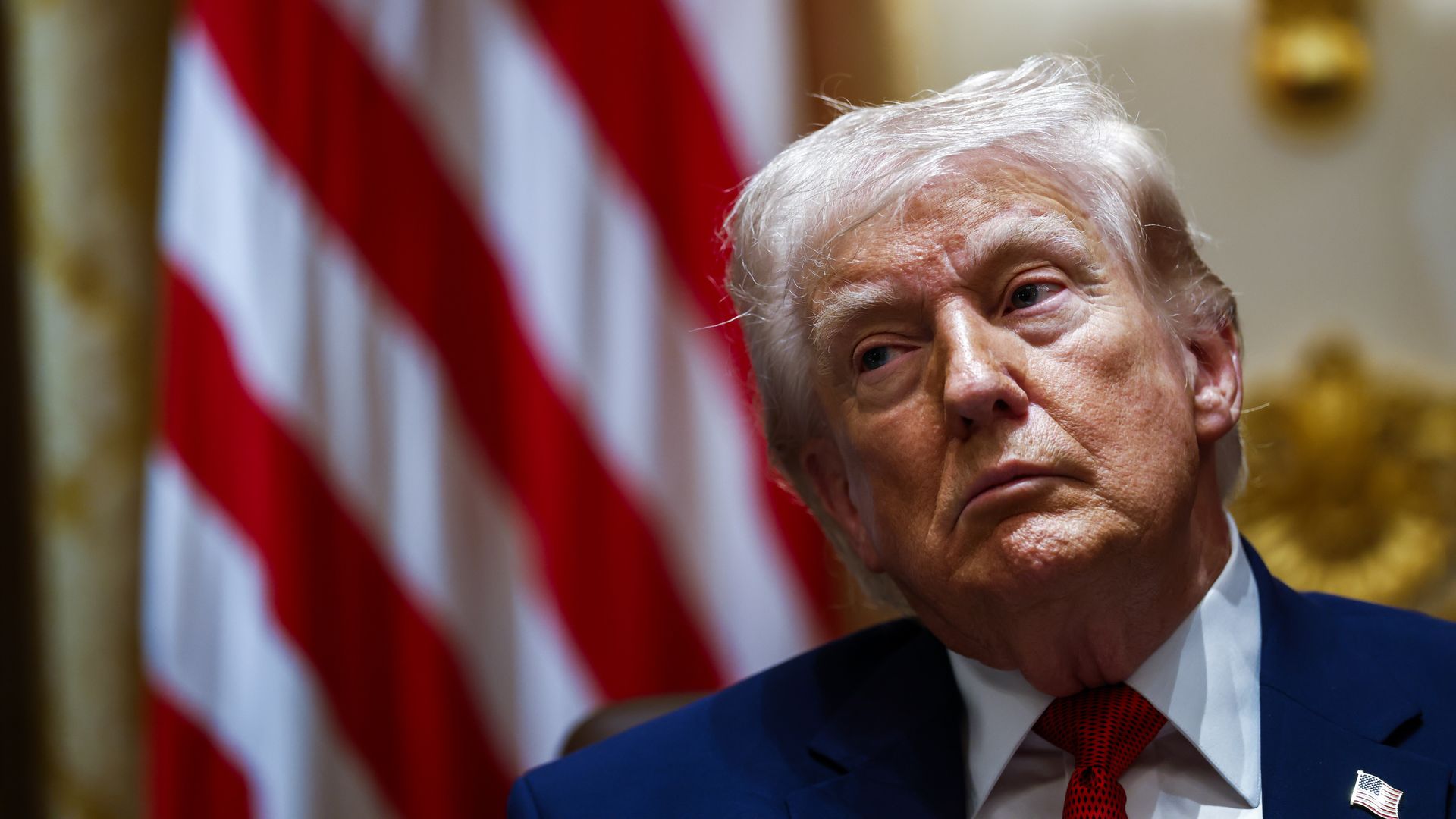 Close-up of President Trump, an older man with white hair in a dark suit and red tie, gazing to the side; blurred American flag and gold decor behind him in a formal setting.