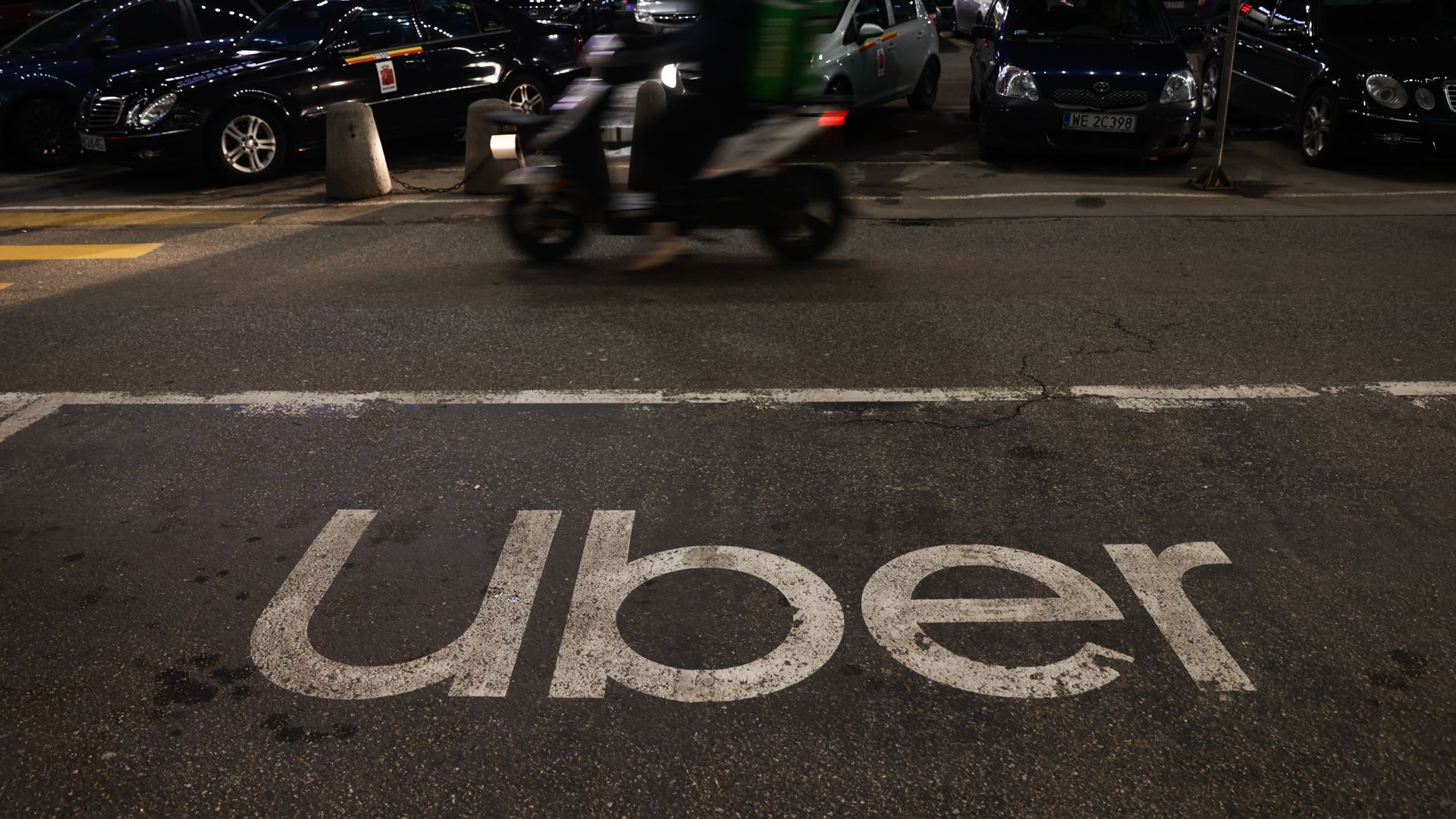 An Uber logo written on the blacktop pavement of a parking lot with a motorcycle zooming past in the background.