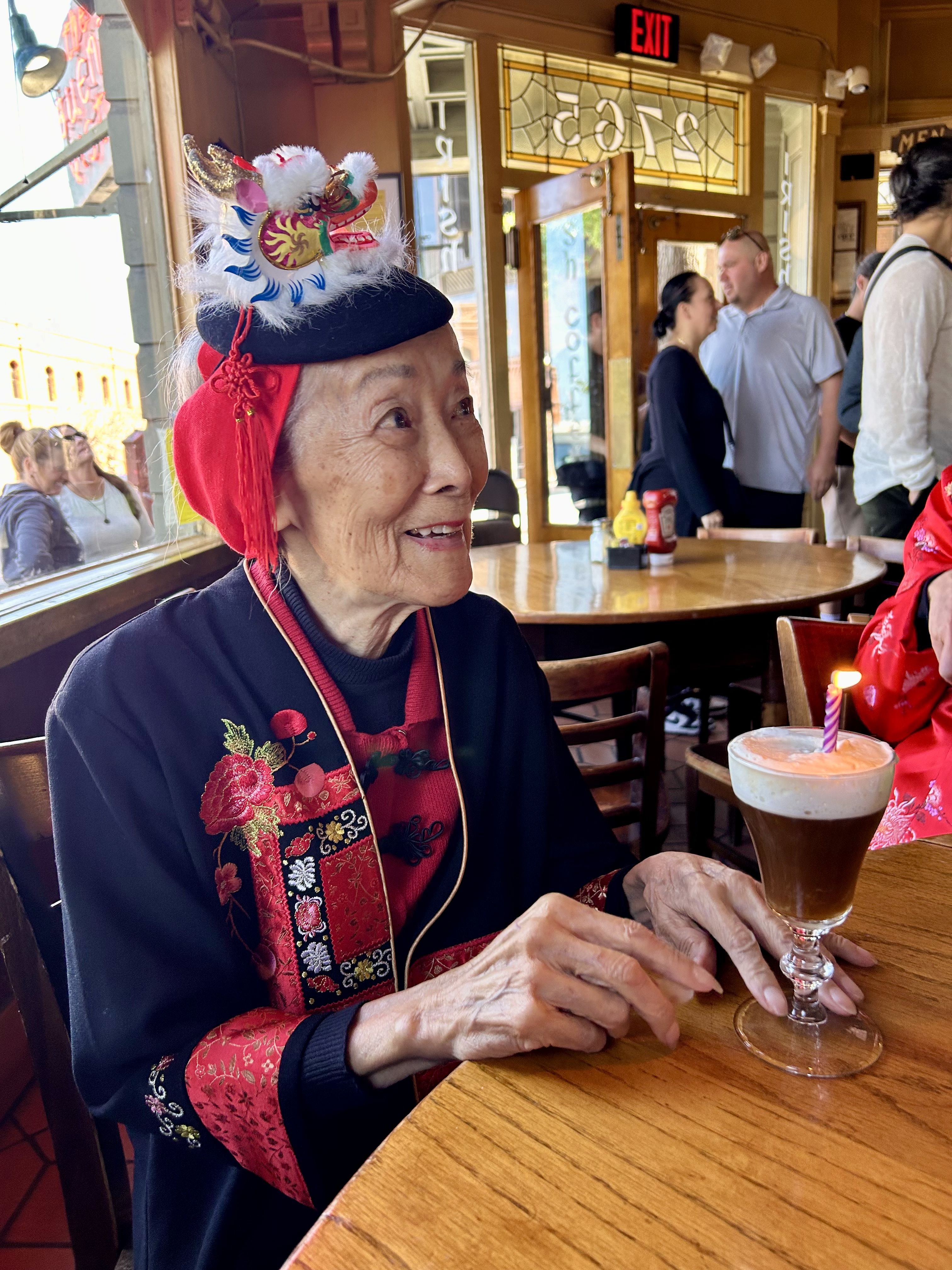 An elderly Asian woman wearing a festive black and red outfit and a decorative dragon hat with white feathers sits at a wooden table in a cafe, smiling, near a tall beer glass with a candle.