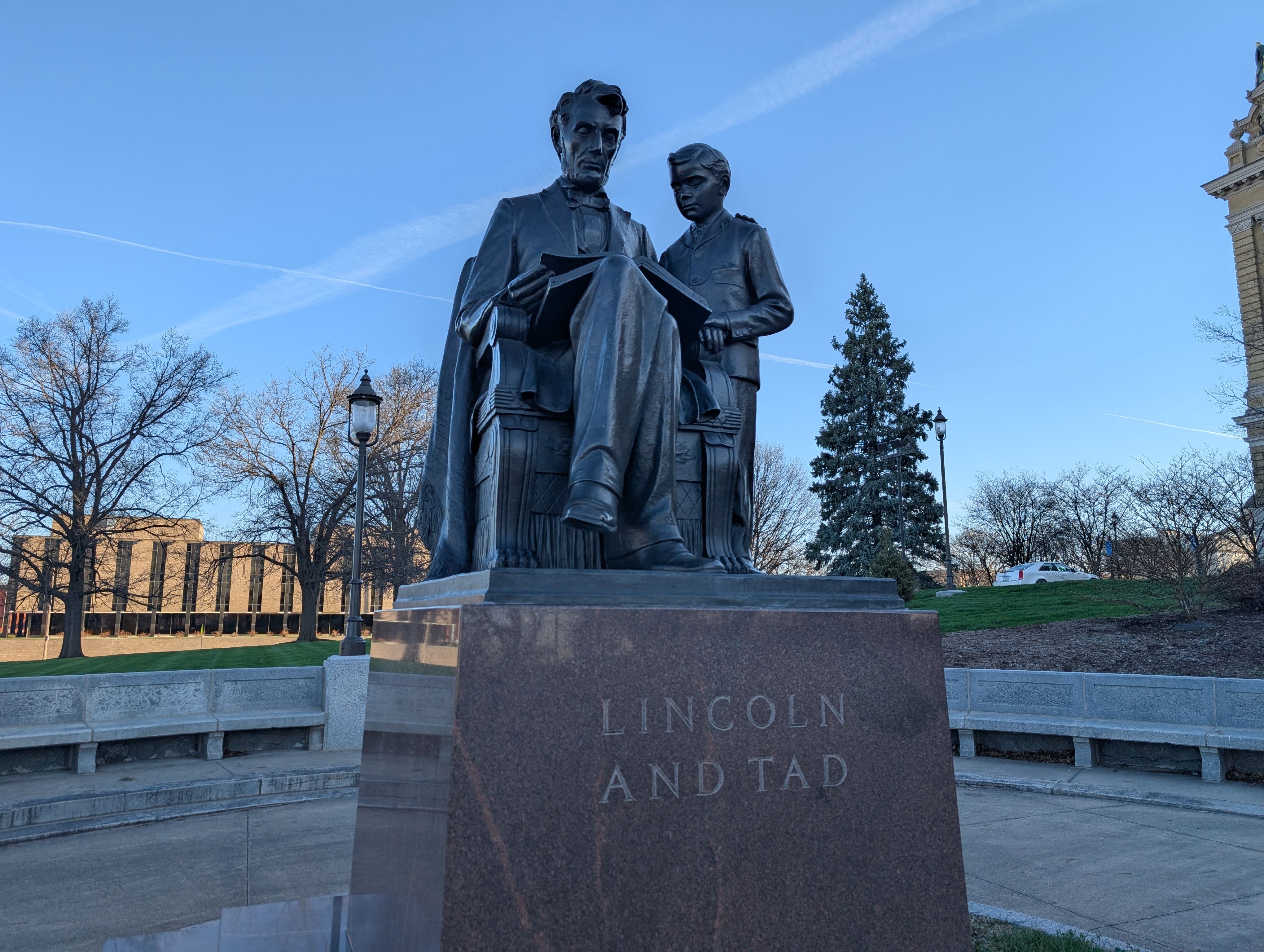 Bronze statue of Abraham Lincoln seated with a boy beside him, reading a book. Pedestal reads "LINCOLN AND TAD" in a plaza under a clear blue sky.