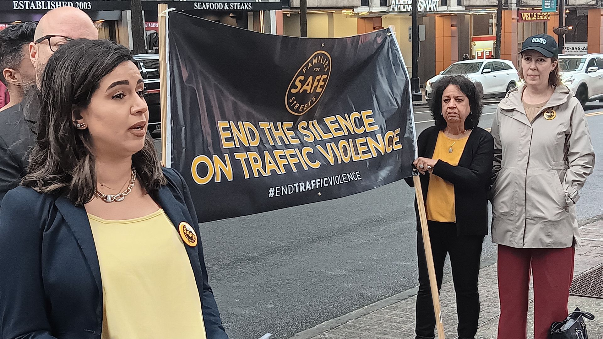 A woman in a yellow top and navy blazer speaks holding papers, beside two women holding a banner that reads "End the Silence on Traffic Violence" on a city street.