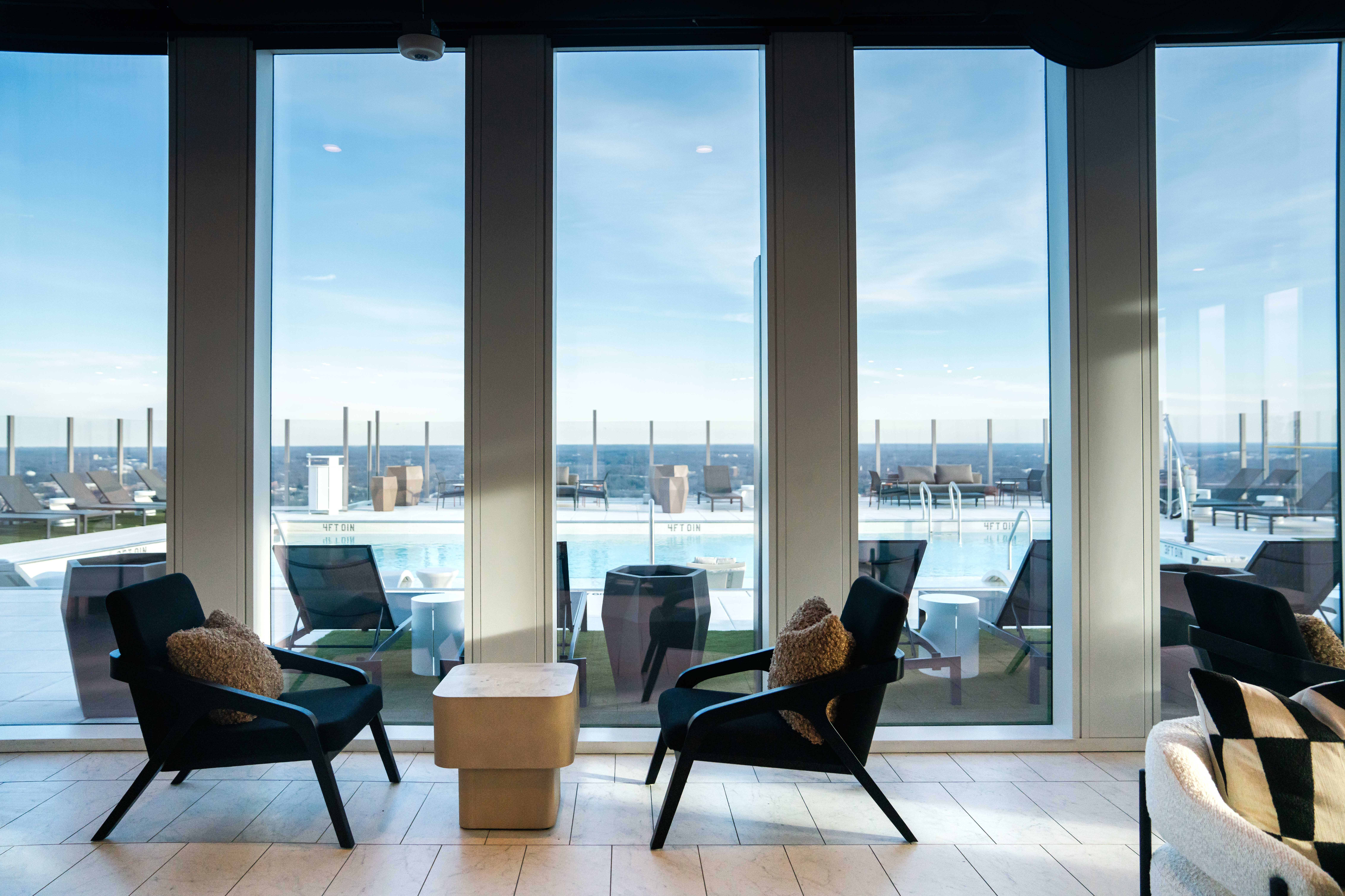 Room interior with two black armchairs with brown cushions and a small beige table between them, facing large windows overlooking an outdoor pool and clear blue sky.