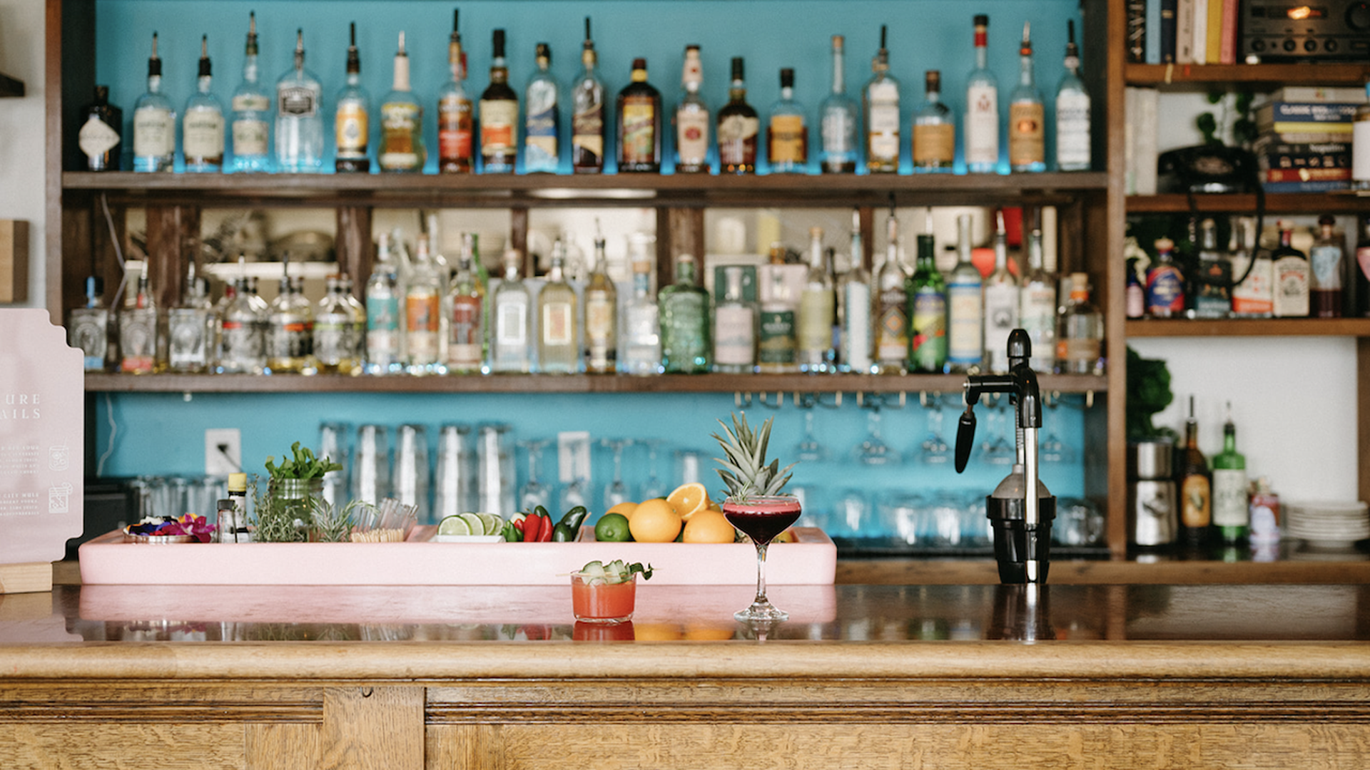 Teal-walled bar with shelves full of bottles. A pink tray holds pineapple, citrus, and herbs on the counter. Two cocktails sit nearby—a short red drink and a dark red coupe; a black siphon stands to the right.