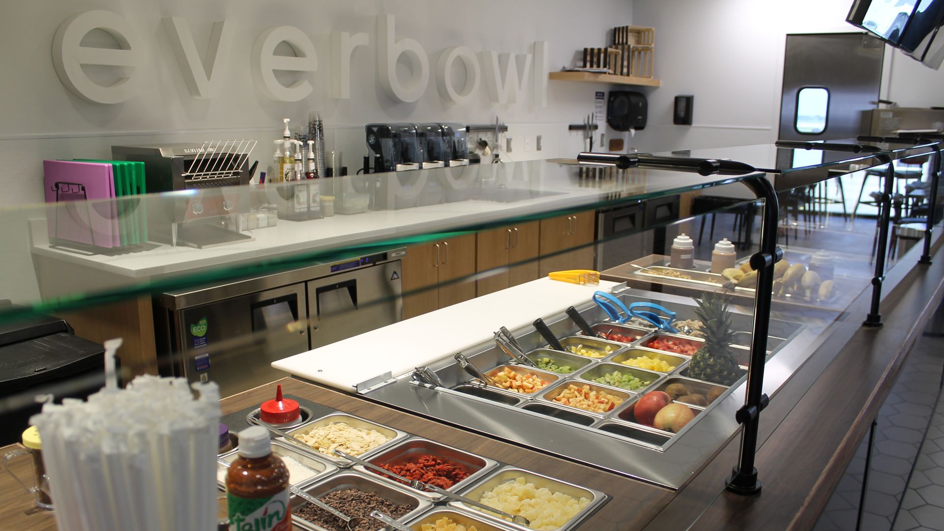 Modern salad bar with a glass sneeze guard, metal trays of diced fruit and toppings, and a bright counter. Large white 3D letters spell "everbowl" on a pale wall behind.