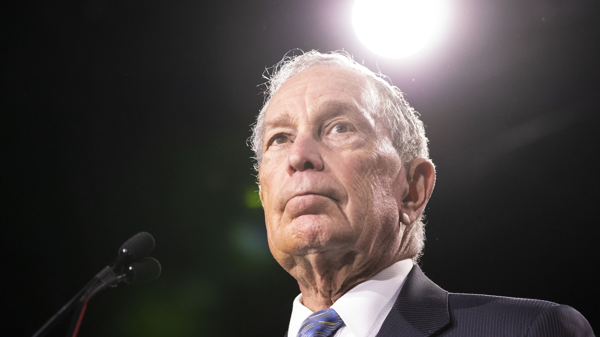 In this image, Bloomberg stands in a suit in front of a microphone.