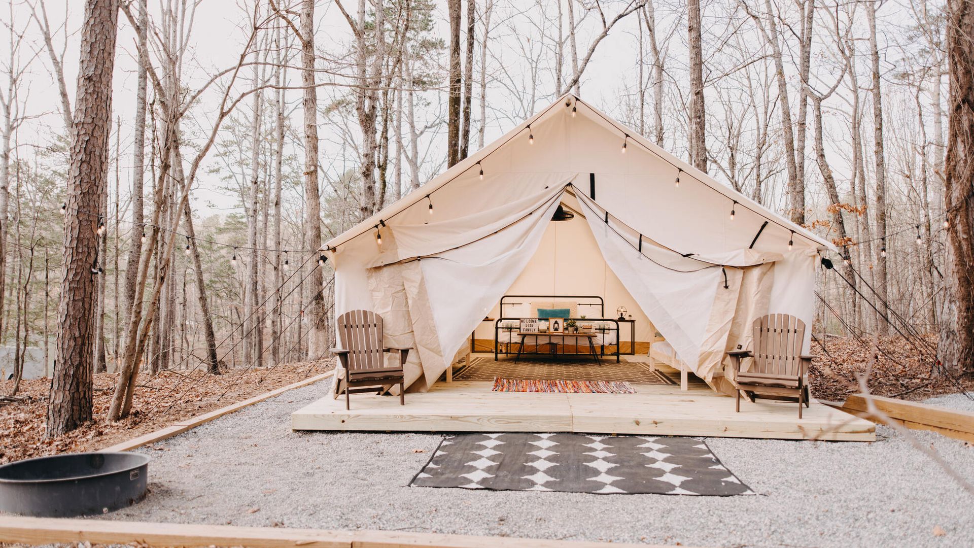 White canvas tent with open front showing two beds and black metal frame, surrounded by bare trees, two wooden chairs, string lights, and a fire pit on gravel