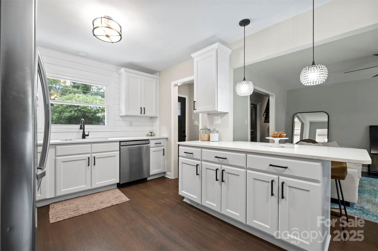 Bright kitchen with white cabinets, dark handles, stainless steel appliances, dark wood floor, window over sink, and two hanging pendant lights over white island countertop.