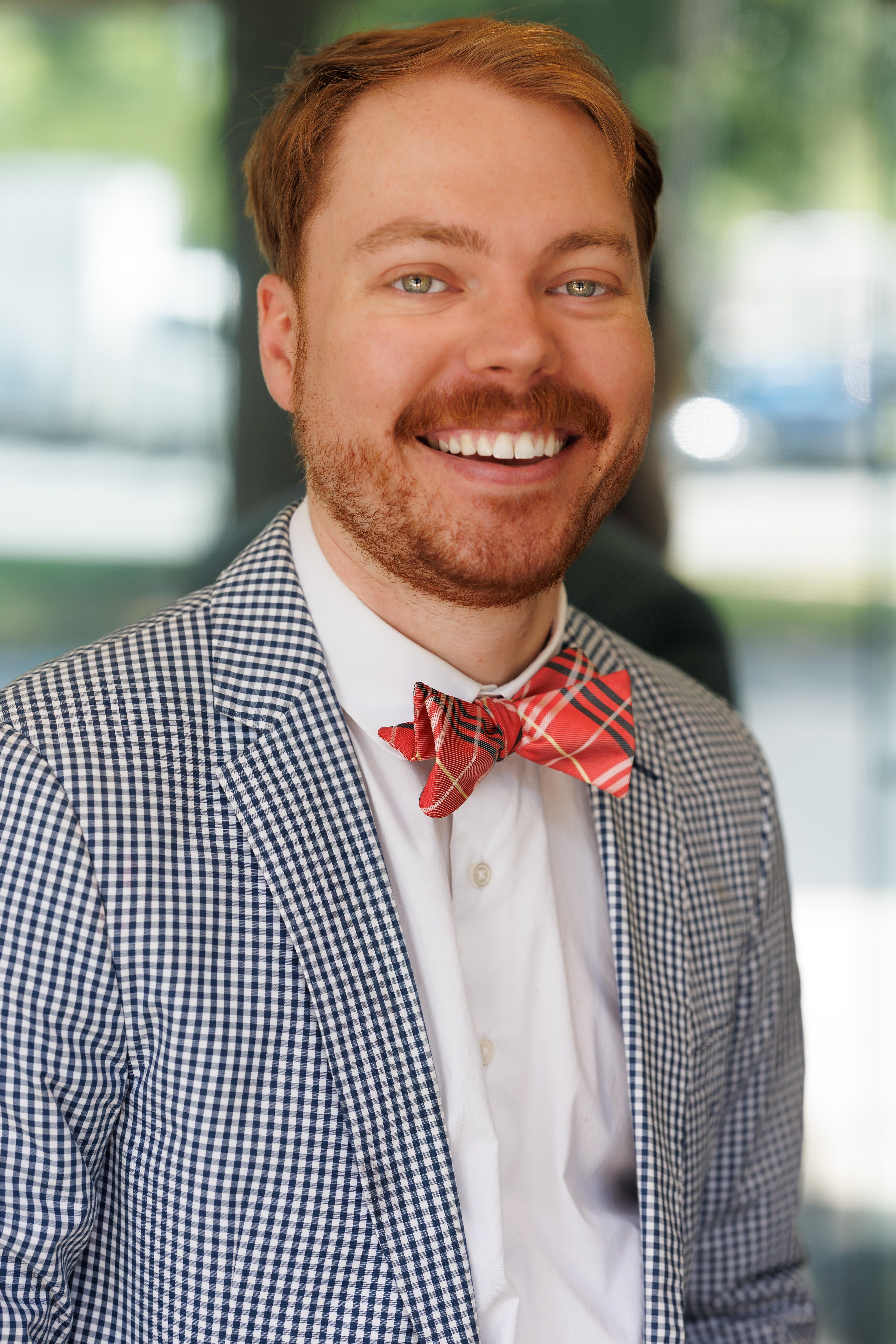 Close-up of a smiling man with light brown hair and a reddish beard, wearing a blue-and-white checkered blazer and a red plaid bow tie, with a softly blurred background.
