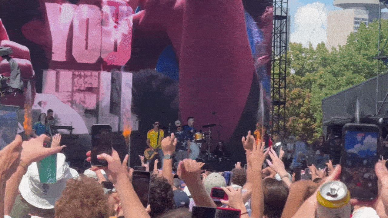 Outdoor concert crowd with raised hands facing a stage. Band members play on a pink-and-purple backdrop; a guitarist in a yellow shirt, drummer, large screen on left; audience holds phones and drinks.