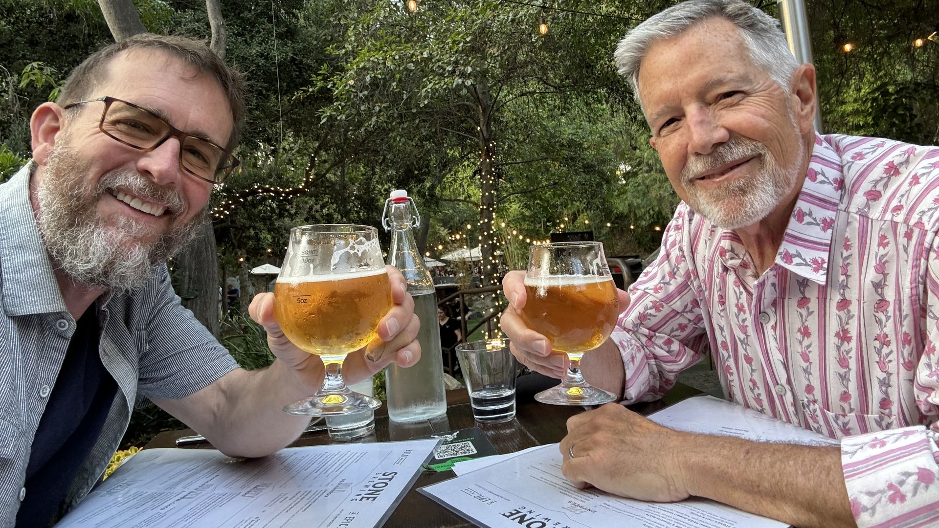 Two men toast amber beers at an outdoor garden restaurant, smiling at the camera. One wears a pink striped shirt, the other a blue shirt. String lights, trees, and a patio heater in the background.