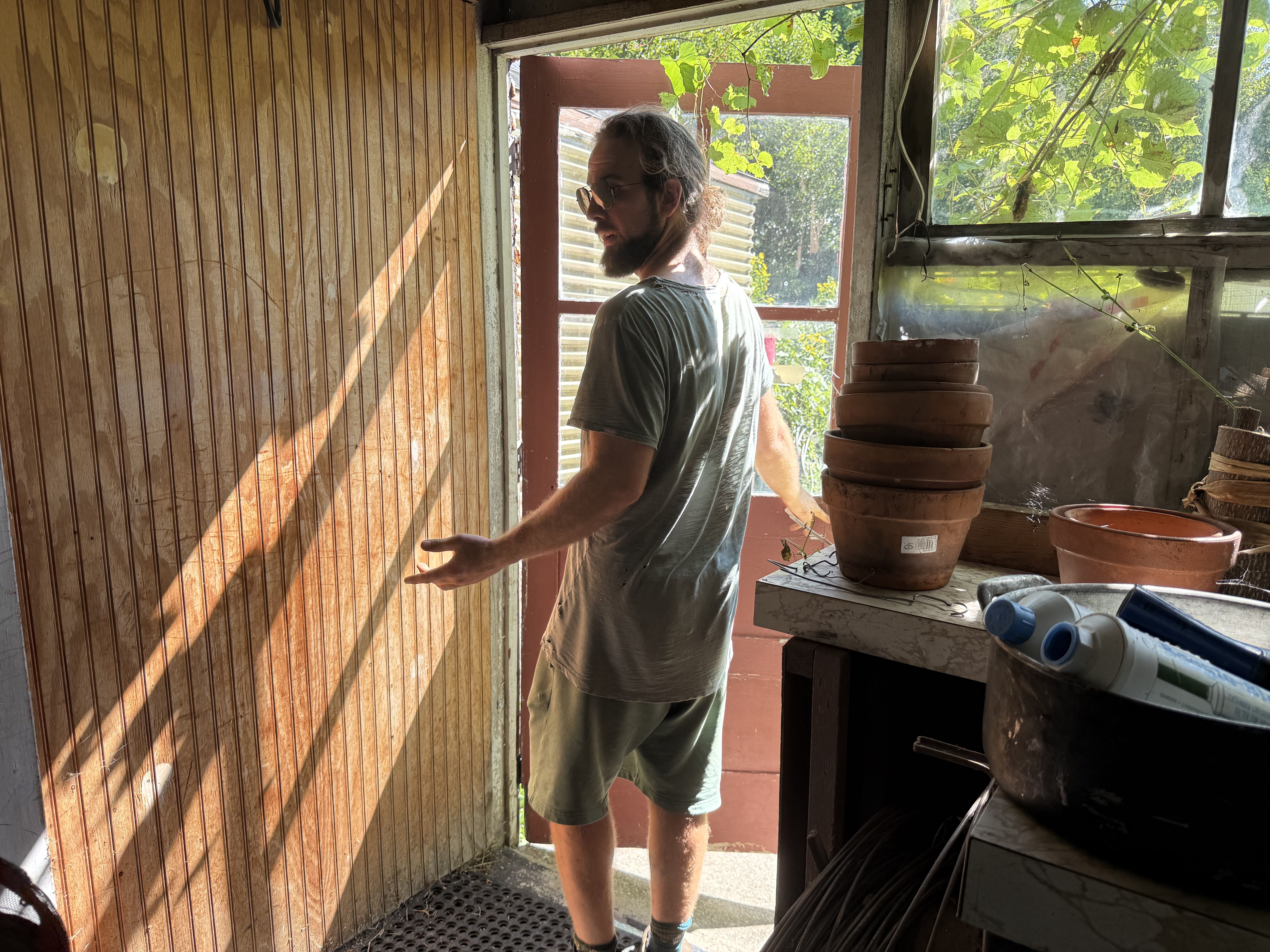 A bearded man with glasses stands in a doorway of a wooden shed, looking to the left. He wears a gray shirt and shorts; stacked clay pots sit on a table, with sunlight and green vines outside.