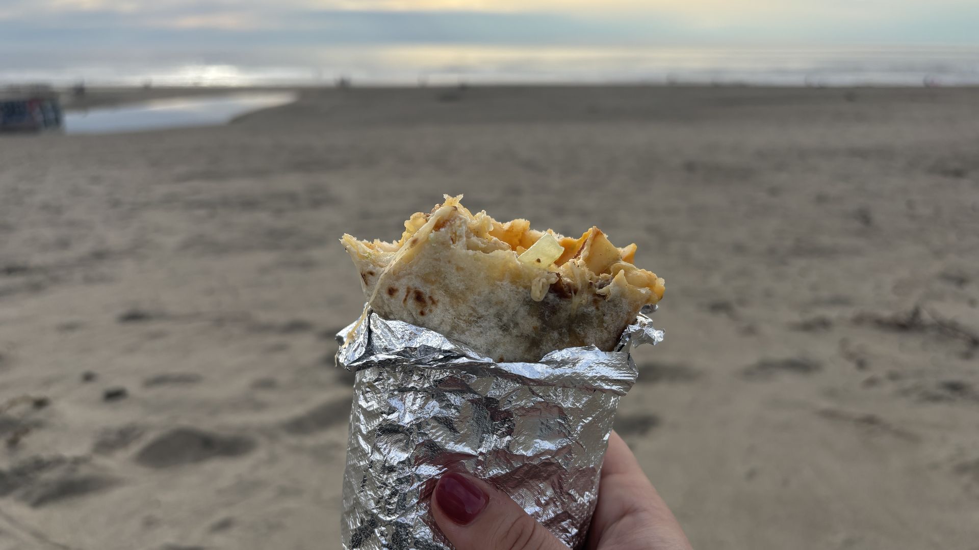 Hand with red nails holds a foil-wrapped burrito on a sandy beach; cheese peeks from the tortilla as the sea and a pale sunset glow on the horizon.