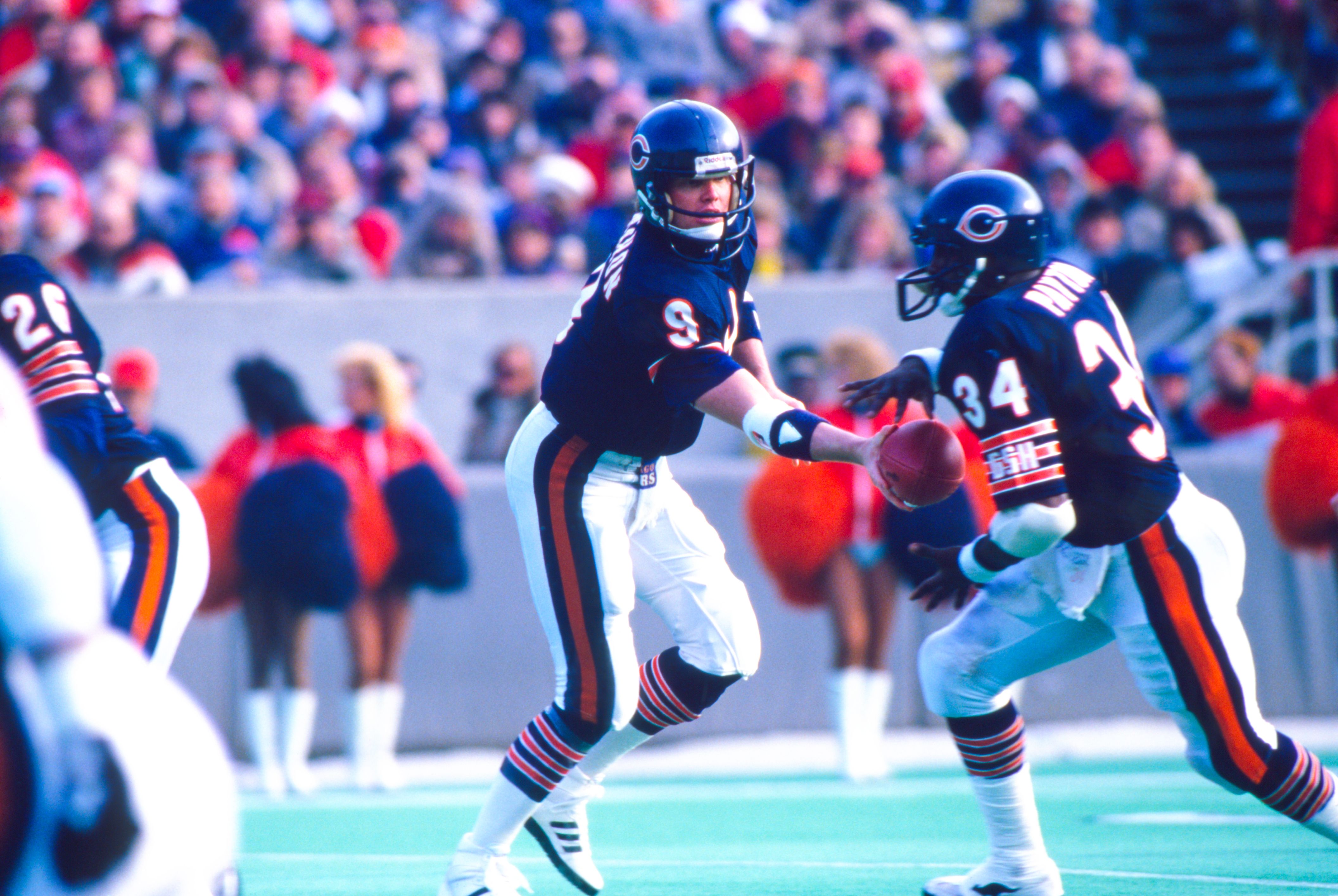 Chicago Bears players in navy blue jerseys and white pants executing a handoff during an American football game with a blurred crowd and cheerleaders in orange in the background.
