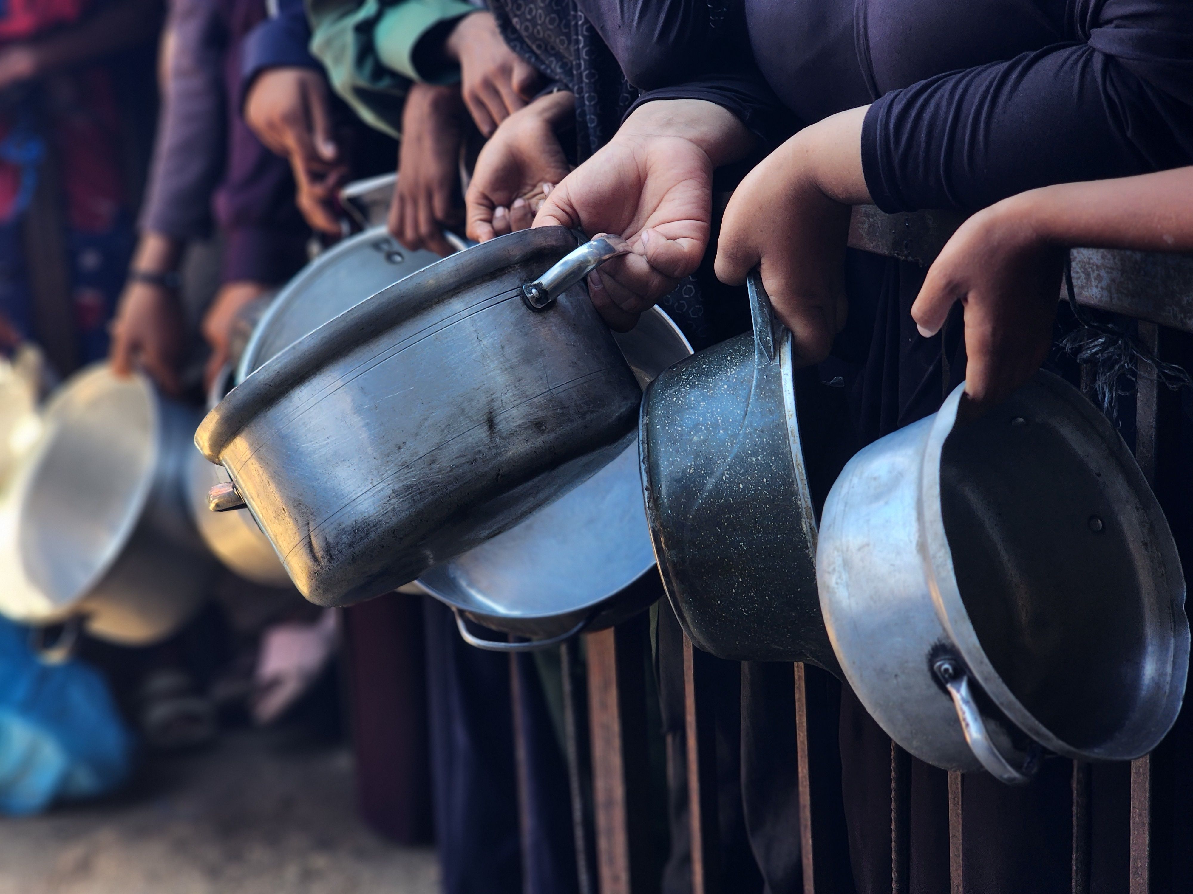 Several people in dark clothing standing in a line holding empty metal pots and pans through a metal railing, likely waiting for food or supplies.