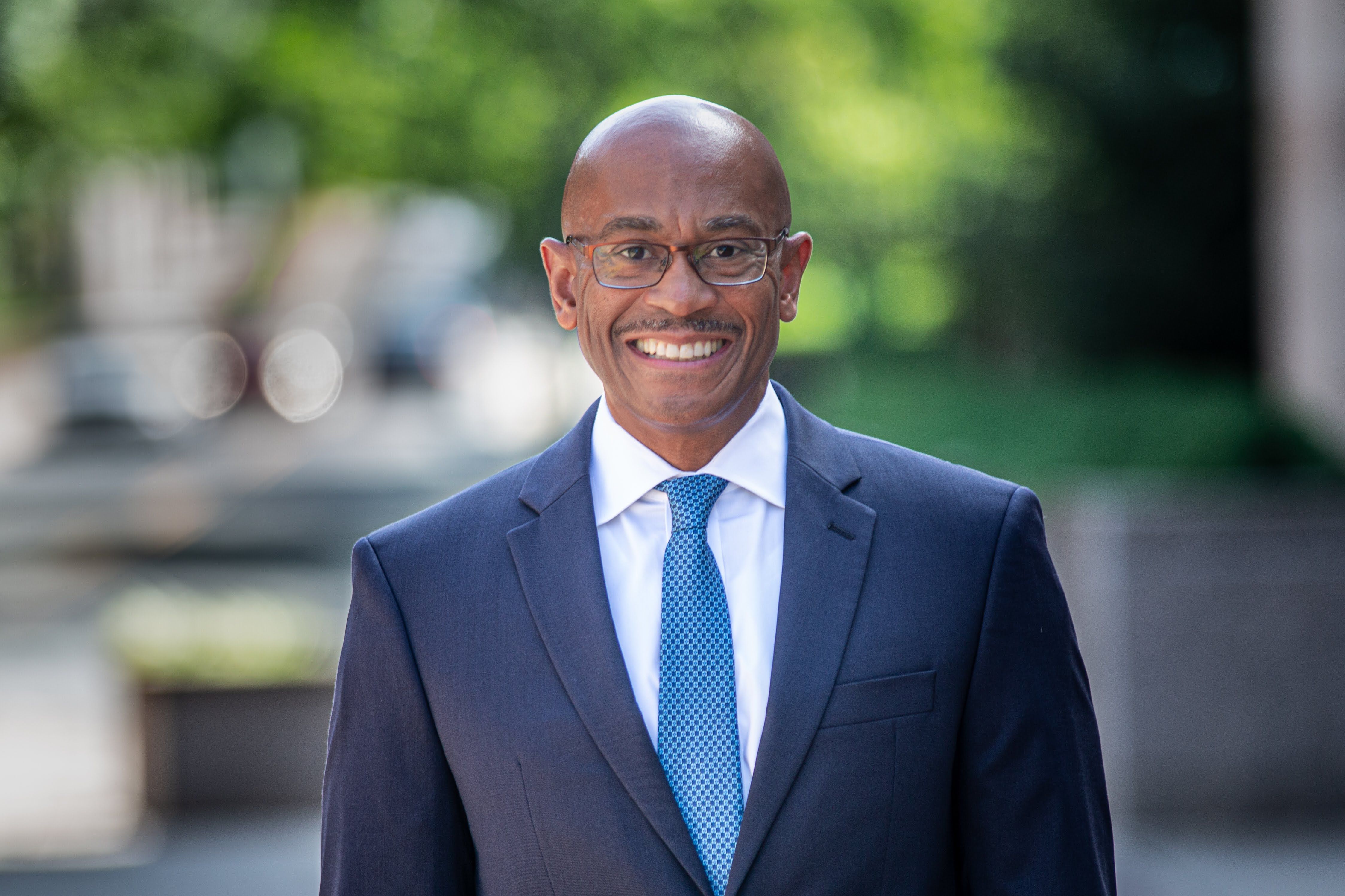 Smiling man wearing glasses, a dark suit, white shirt, and patterned blue tie standing outdoors with blurred green and gray background.