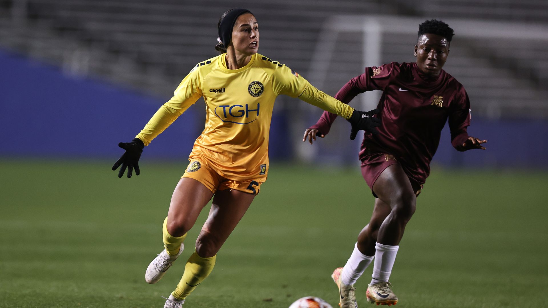 Two female soccer players competing on a field at night; one in yellow kit running with arms outstretched, the other in maroon kit chasing after a soccer ball on the grass.