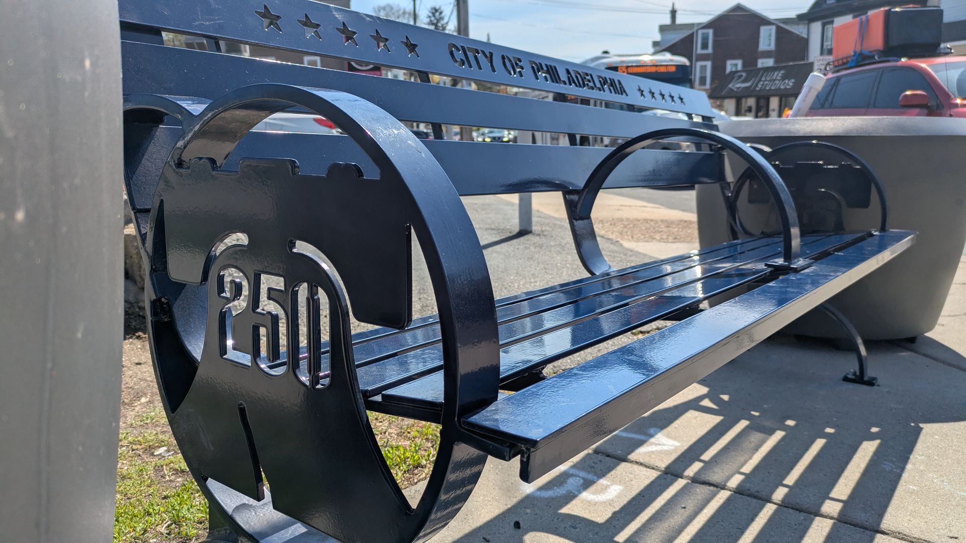 Close-up of a glossy blue metal bench with a backrest that reads City of Philadelphia and star cutouts; a circular end displays 250. Cars and buildings line a sunny street in the background.