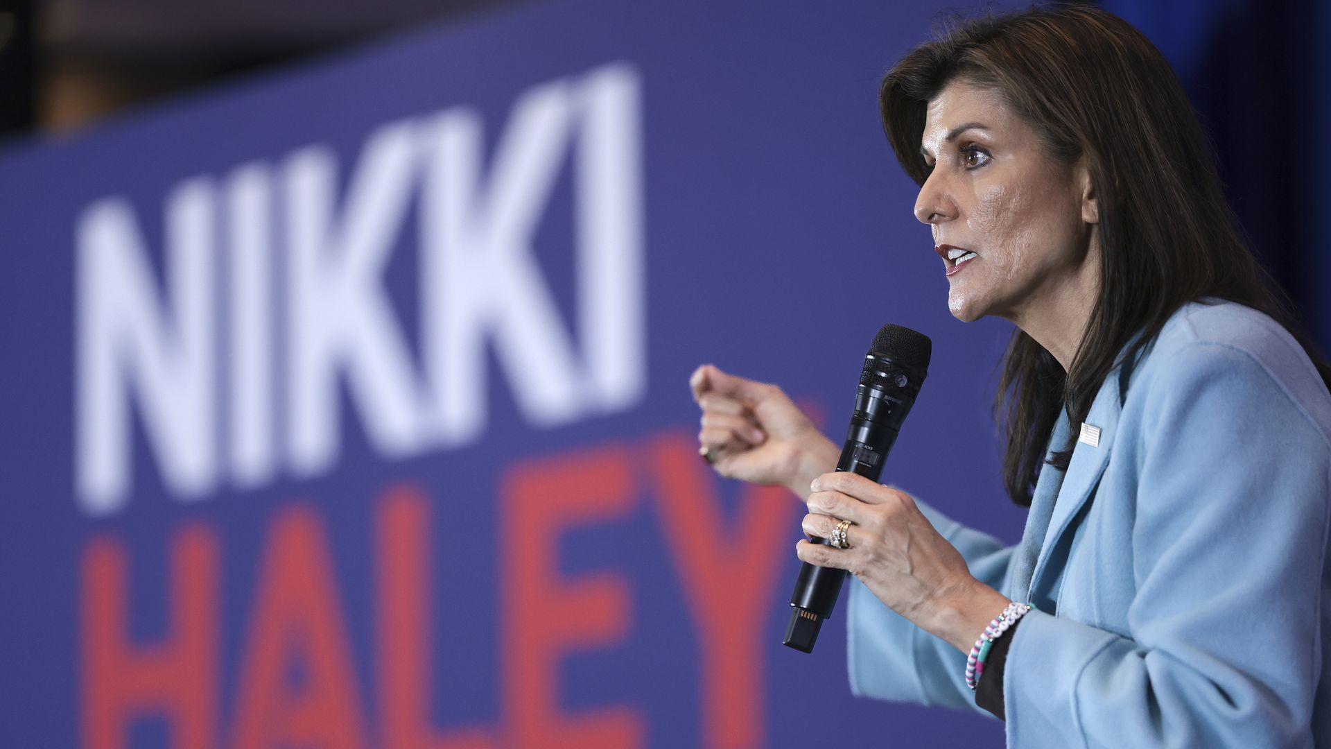 Nikki Haley, wearing a blue jacket, speaks holding a microphone. A large sign in the background says her name in all caps.
