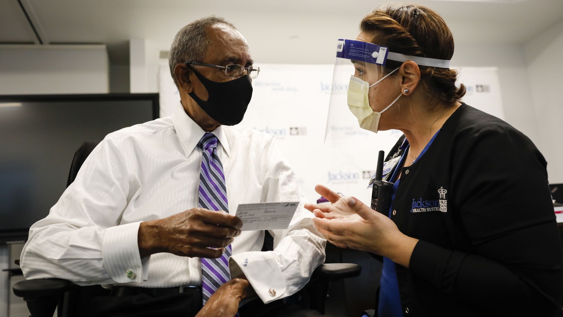 A woman wearing a face shield, face mask and PPE hands a card to a man in a suit and face mask 