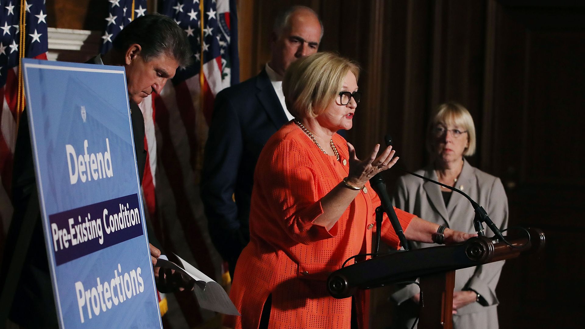 Sen. Claire McCaskill speaking next to a sign that says defend preexisting conditions