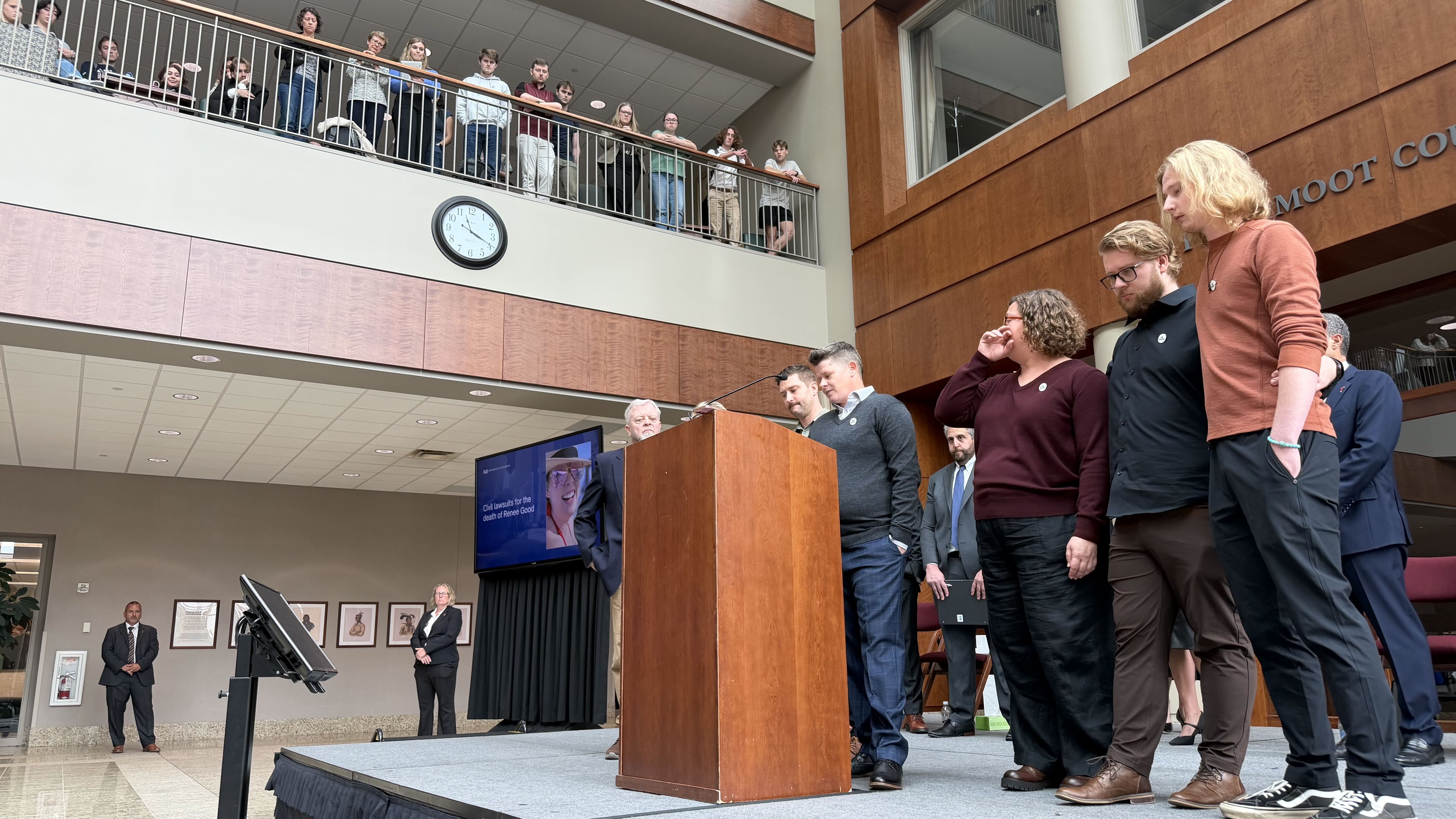 Scene in an atrium: a podium with microphones on stage, several people in smart casual wear; a clock on the wall, brown wood panels, and spectators on a second-floor balcony looking down.