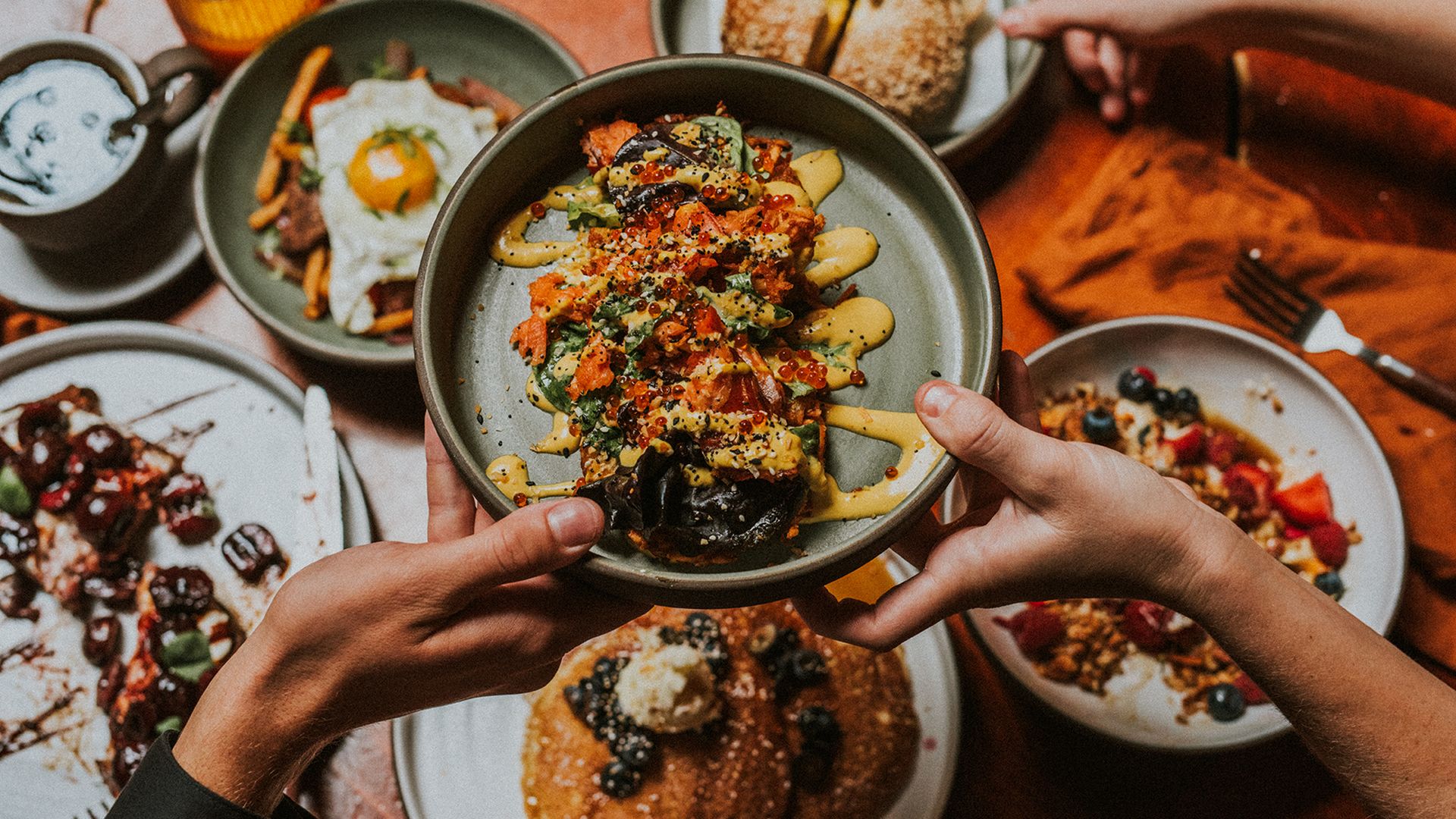 Two hands exchanging a bowl of orange, green, and yellow vegetable dish over a table with pancakes, berry-topped granola, fried egg, chocolate dessert, and a sesame bagel.