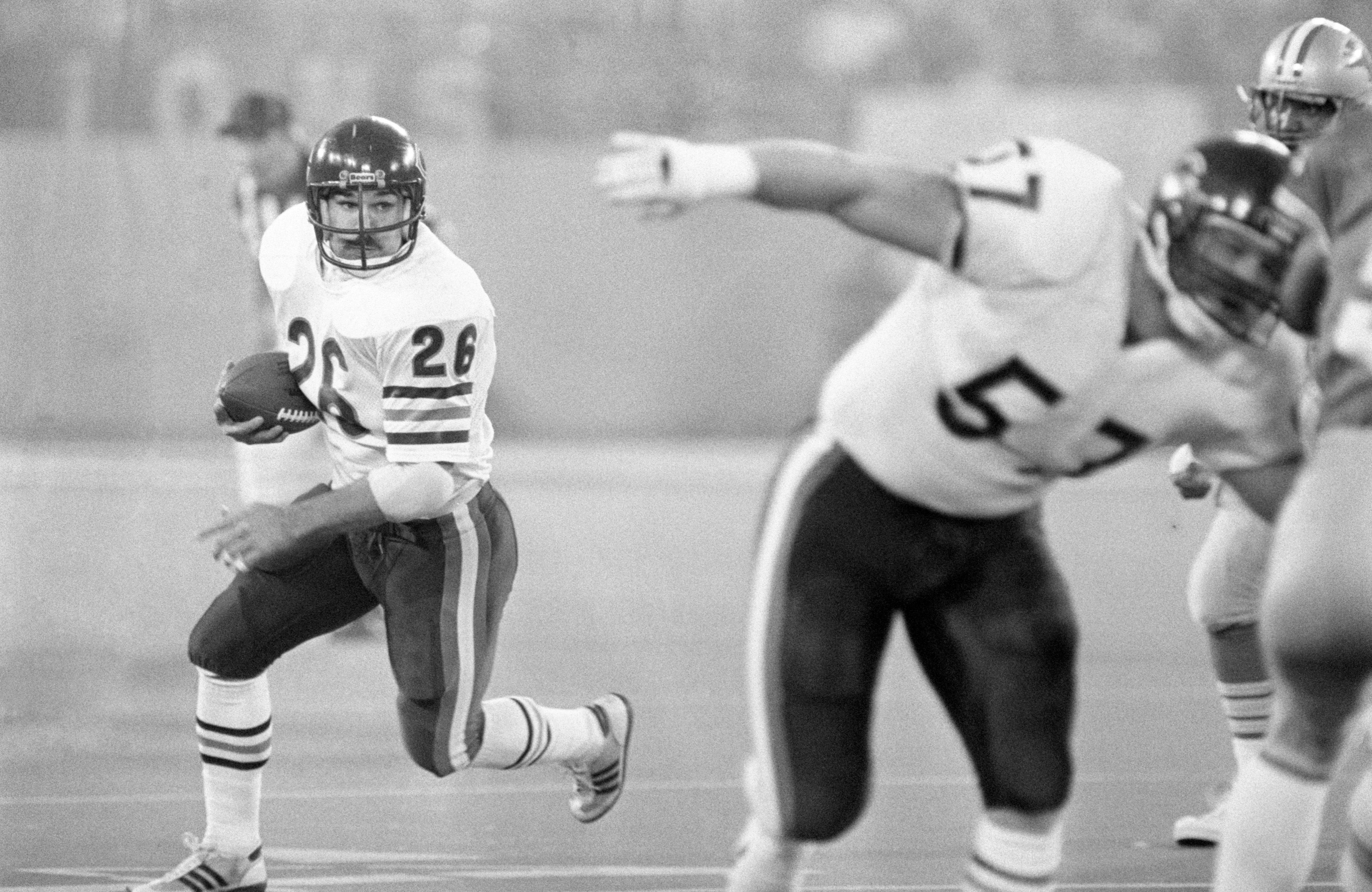 Black and white photo of football player #26 in white jersey running with ball, looking left, as teammate #57 in white jersey blocks opponents during game.