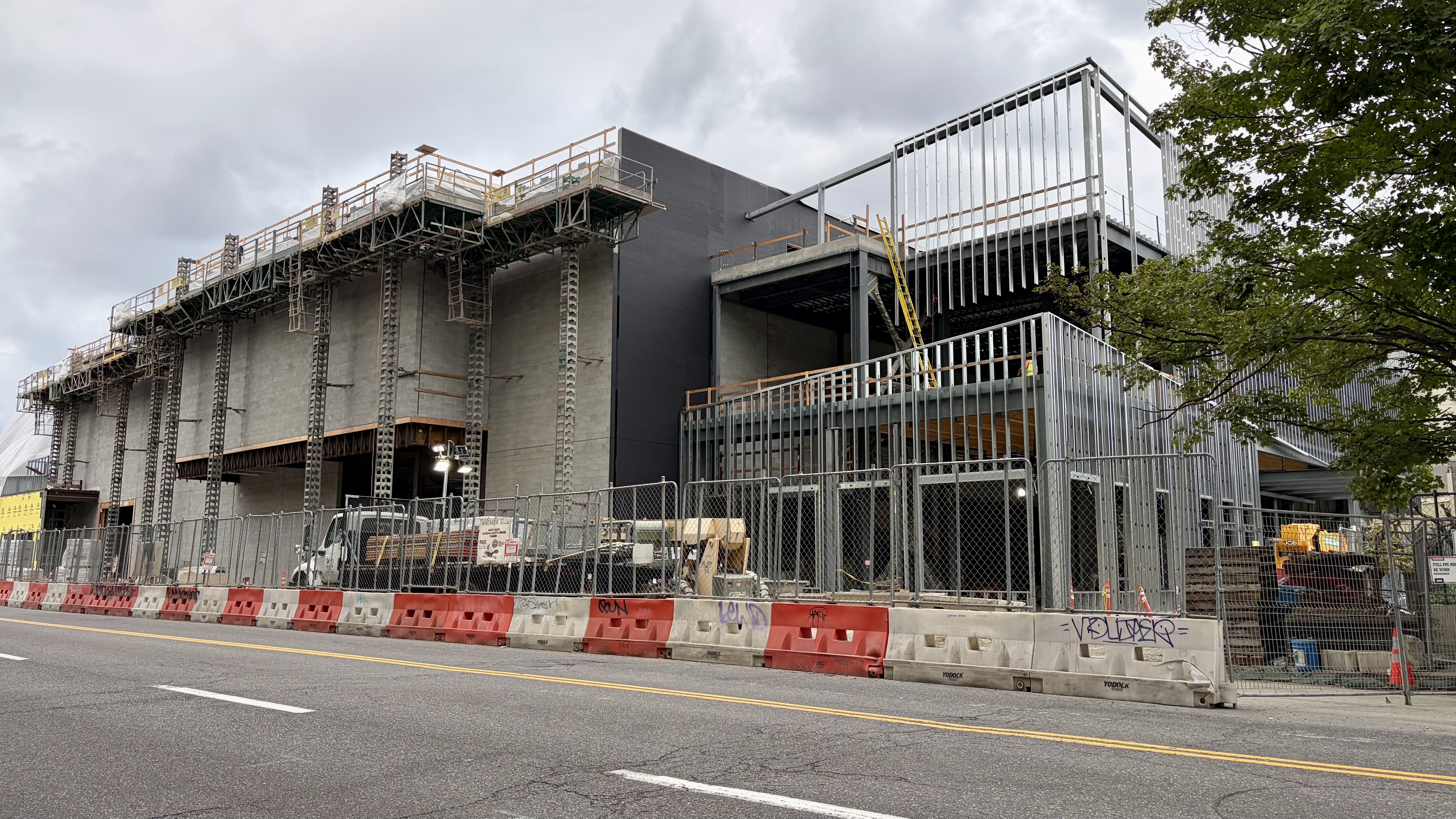 Construction site with a gray concrete building under heavy scaffolding and metal framing; orange barriers line the street, a chain-link fence, ladders, and a cloudy sky with trees on the right.