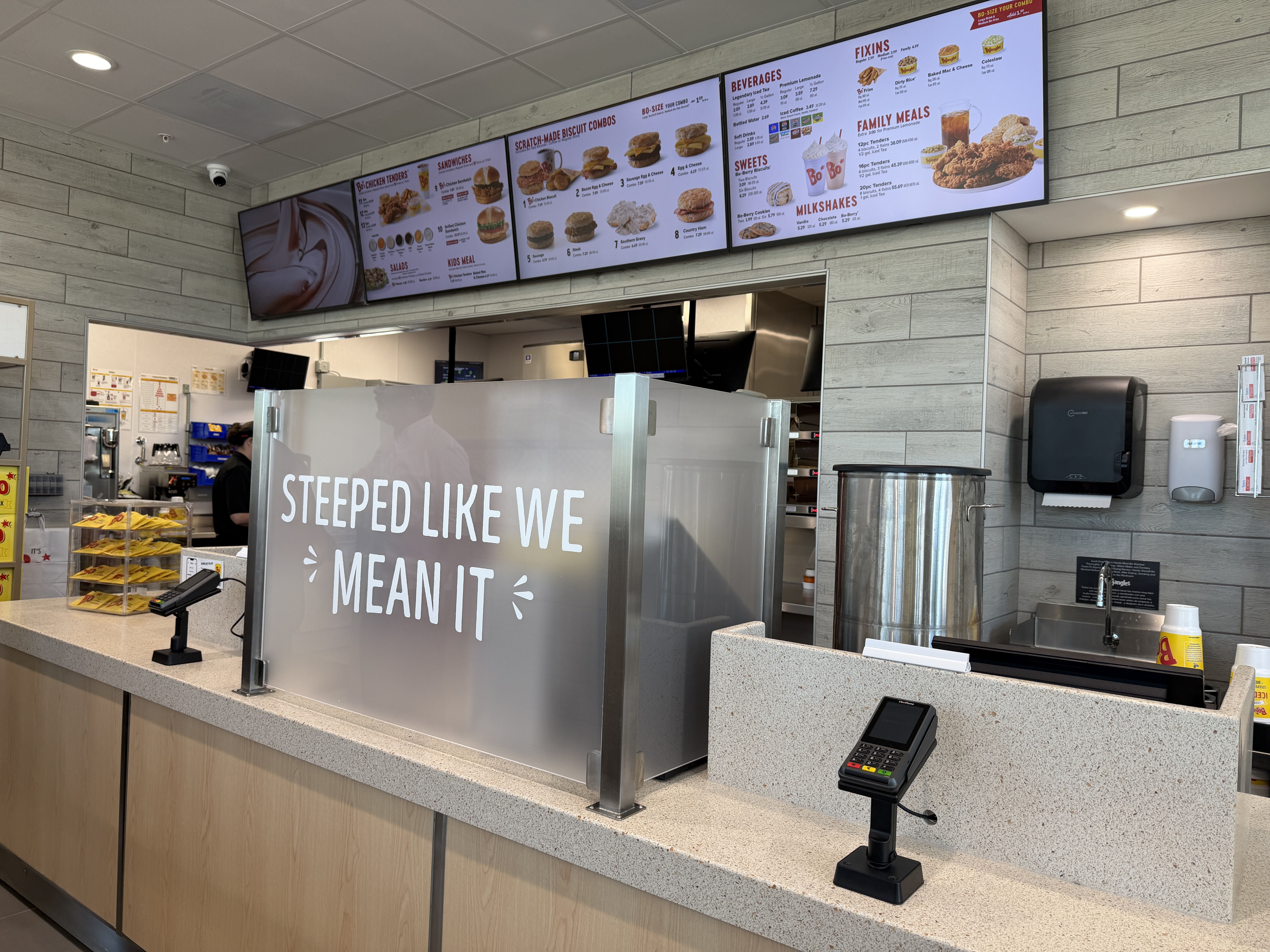 Inside a fast-food restaurant, a long counter with a frosted glass panel reading STEPPED LIKE WE MEAN IT. Overhead menu boards display burgers and beverages; gray wood walls and ceiling lights.