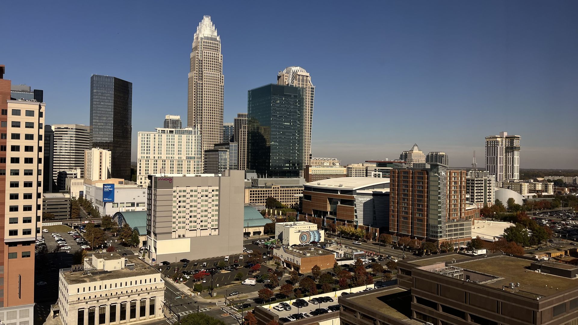 City skyline under a clear blue sky, with tall modern skyscrapers—one central white-crowned tower and glassy blue buildings—surrounded by lower office blocks and a parking lot in the foreground.