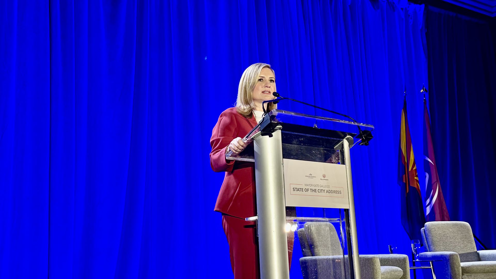 A woman in a red blazer gives a speech at a clear podium on a stage with deep blue curtains, microphones, and flags in the background. A sign on the front of the podium reads "State of the City Address."