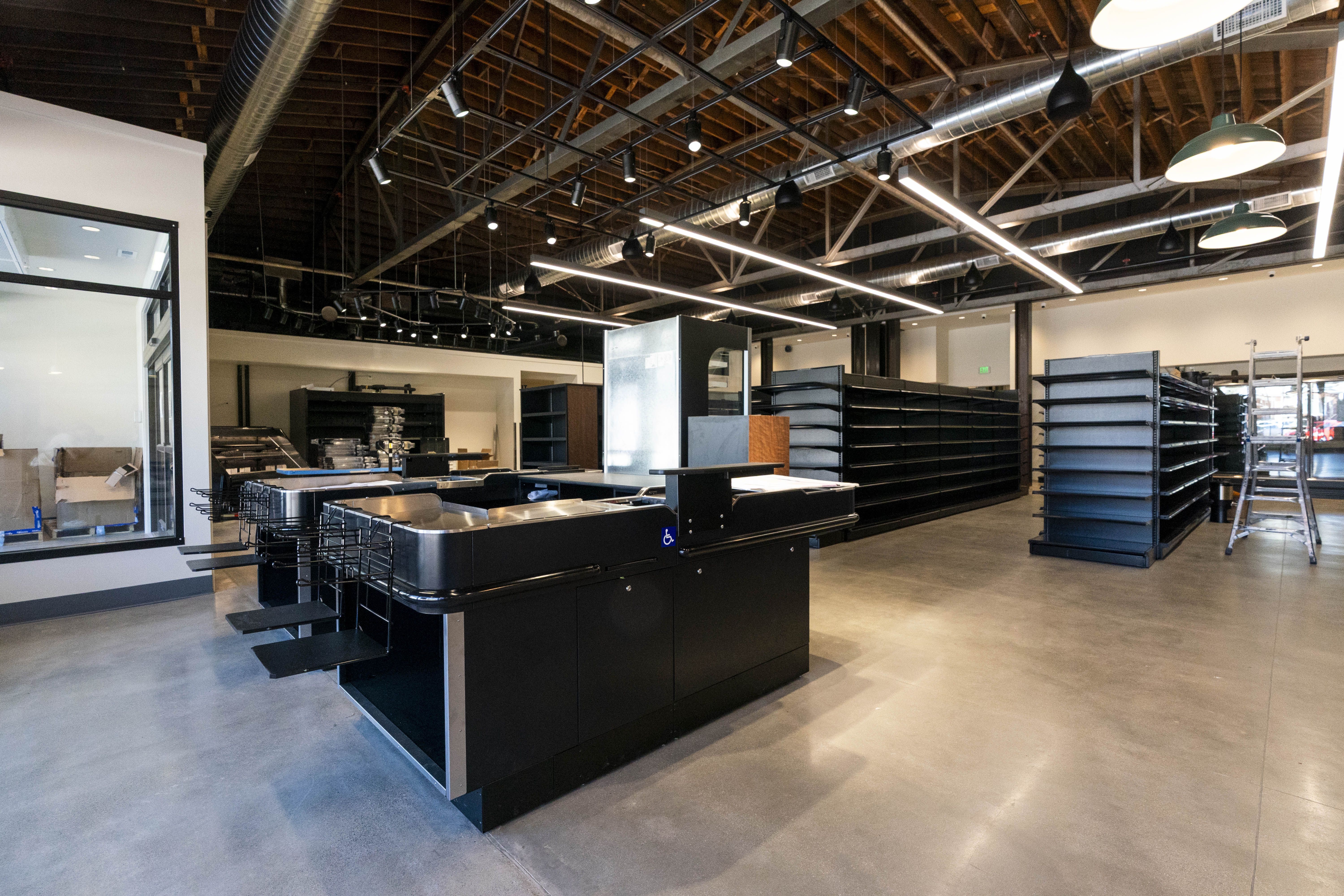 Interior of a modern retail store under construction, with exposed ceiling pipes, track lighting, and rows of black shelving. Checkout counter in foreground; ladder on right; large windows left.