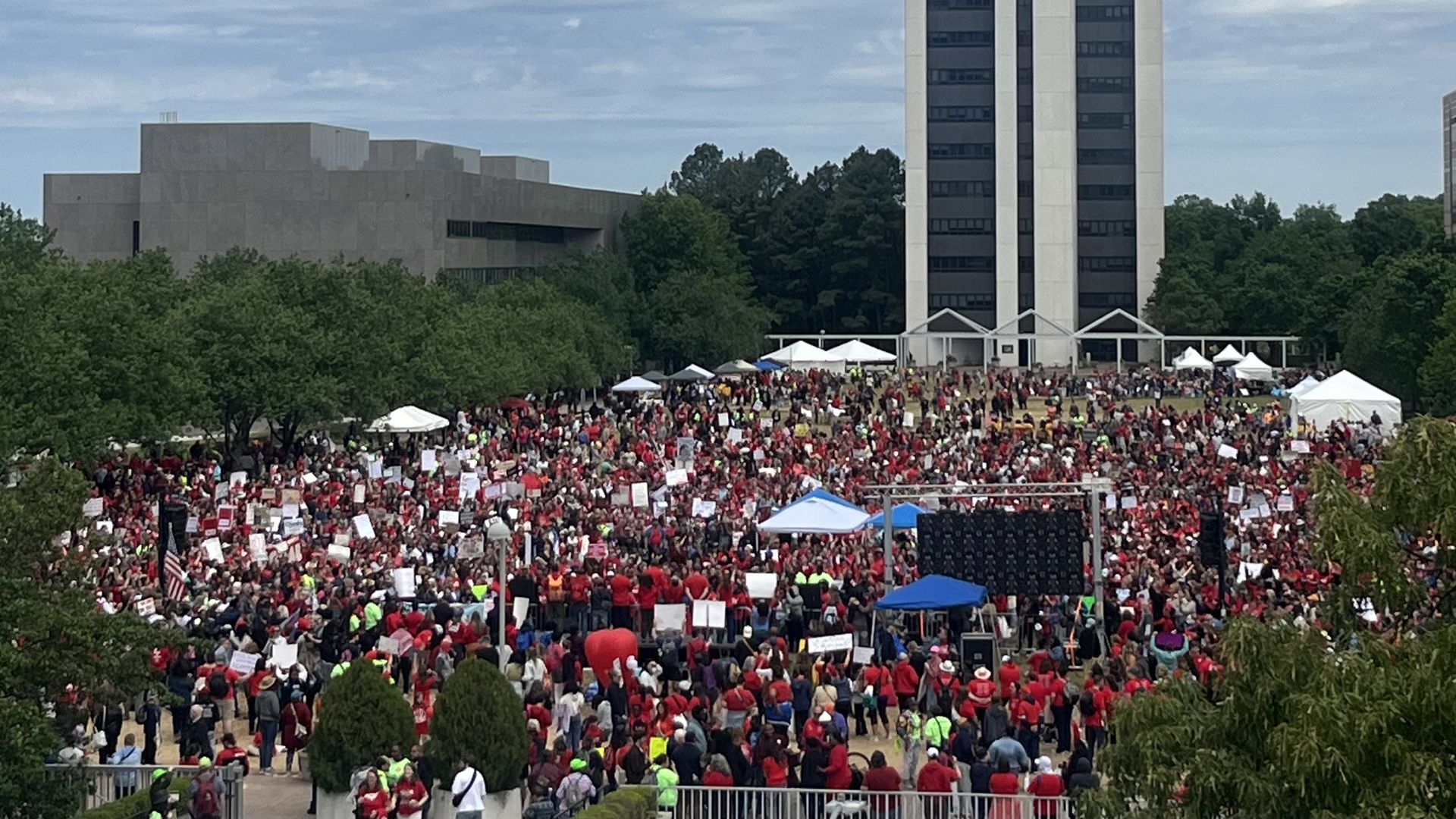 Large outdoor rally with hundreds of people in red shirts holding signs gathered in front of a tall modern gray building. White tents and a stage with screens under a blue, cloudy sky.