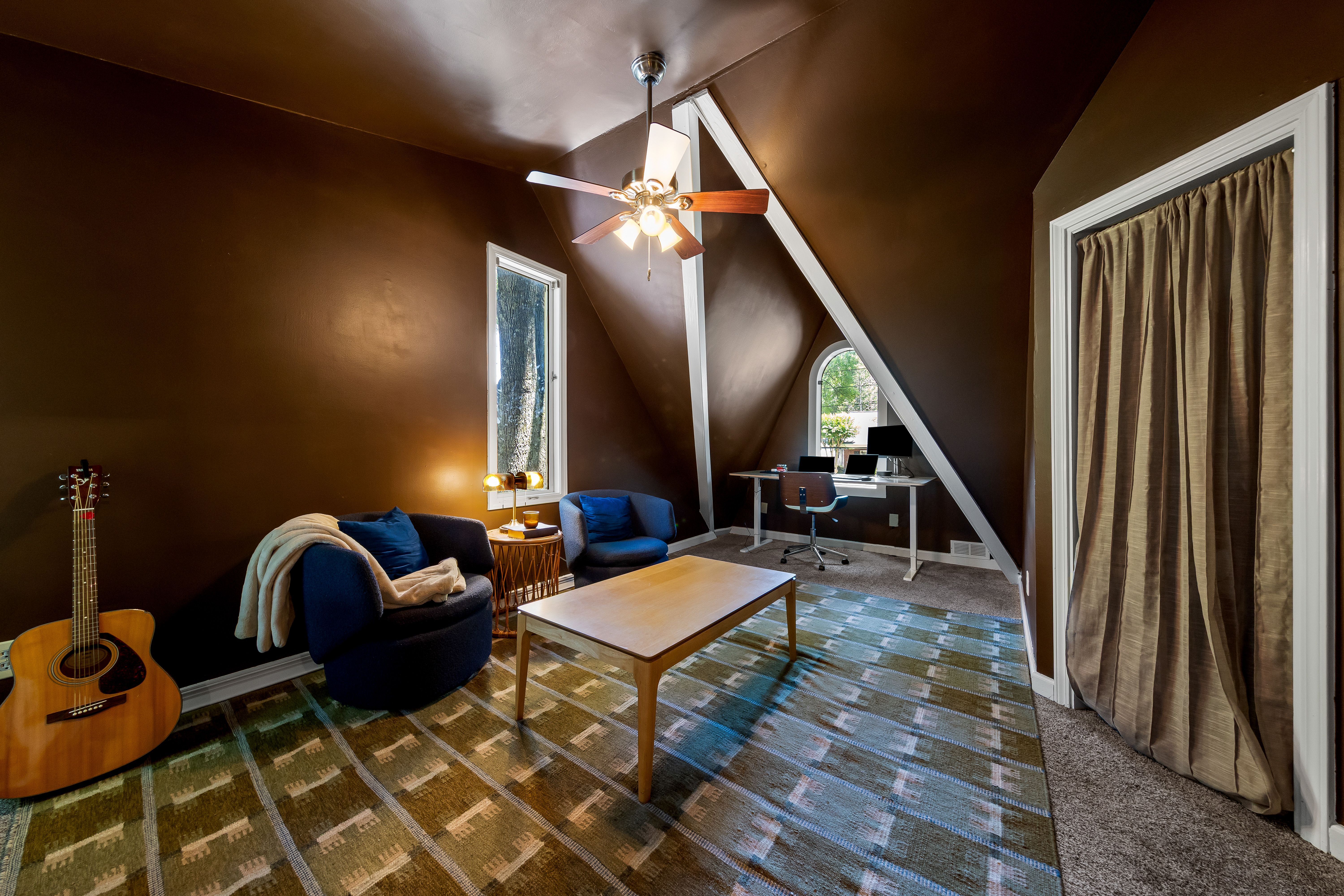 Cozy living room with dark brown walls and a slanted ceiling. A wooden-blade ceiling fan over blue seating; guitar by the couch. Coffee table on a striped rug, with windows and curtains at end.