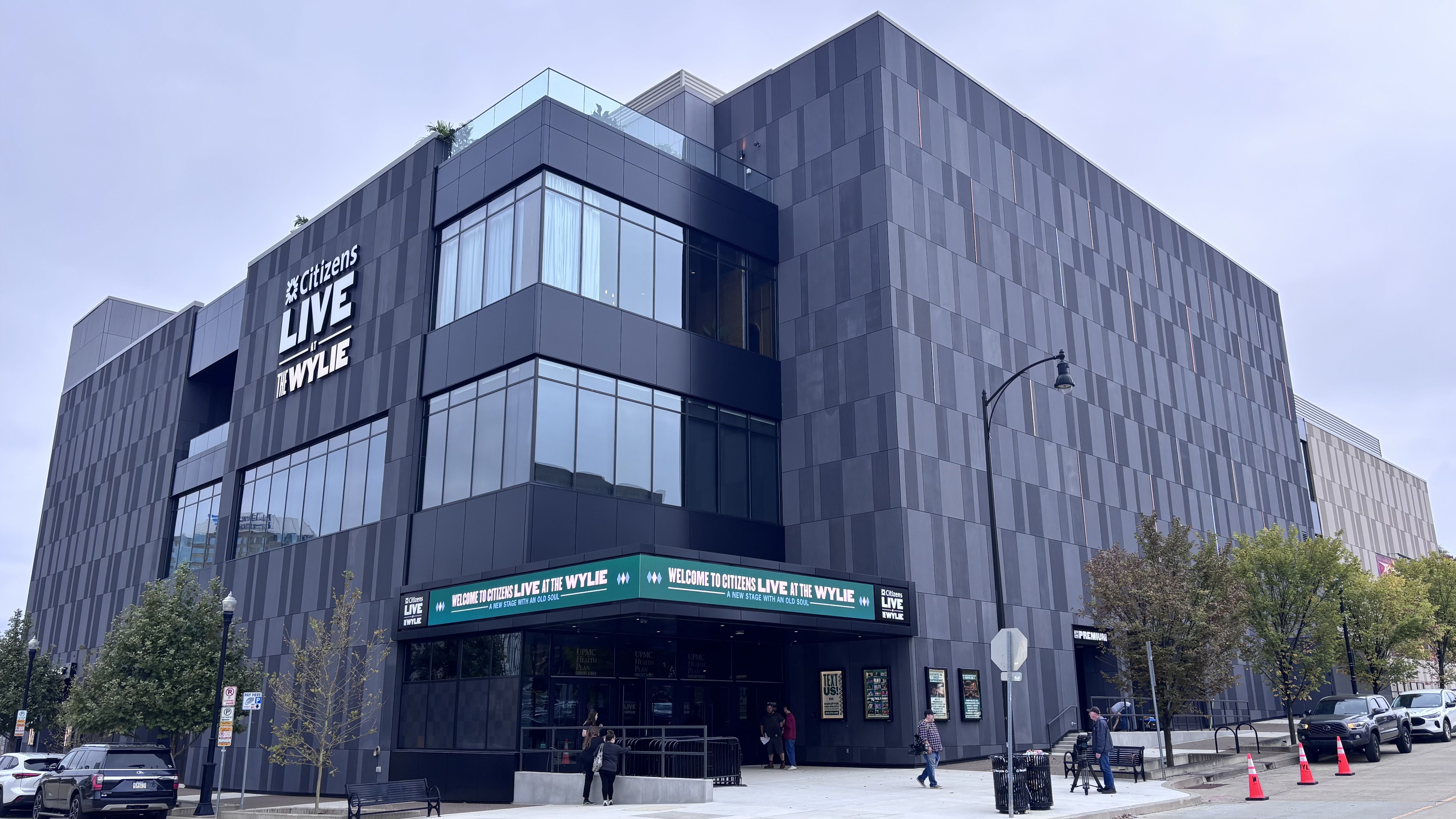 Modern dark gray office building with large glass windows and a glass-roof terrace. Prominent sign reads "Citizens LIVE AT THE WYLYE" on the facade; street with cars and pedestrians.