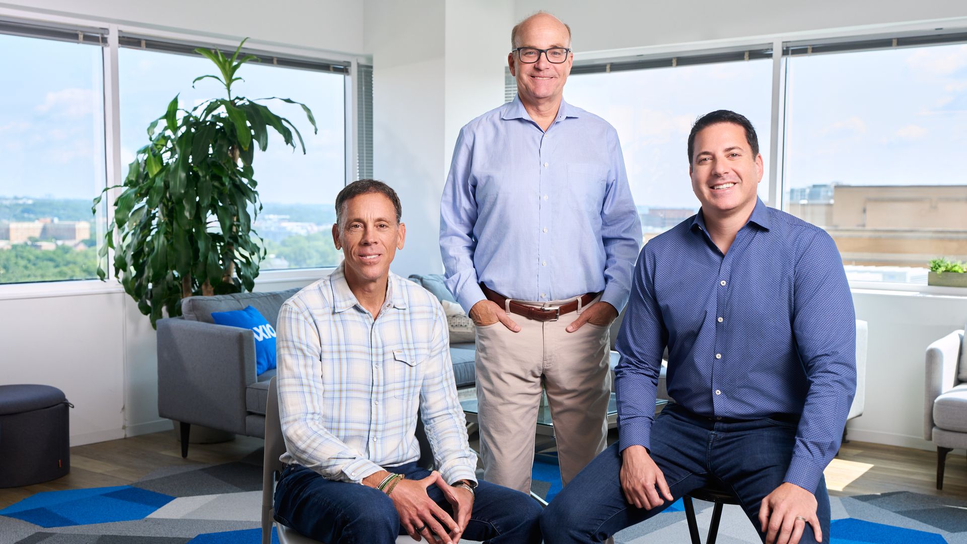Three men in an office with large windows and a plant. One seated in a plaid shirt, one standing center in a light blue shirt and glasses, the other seated right in a dark blue shirt, all smiling.